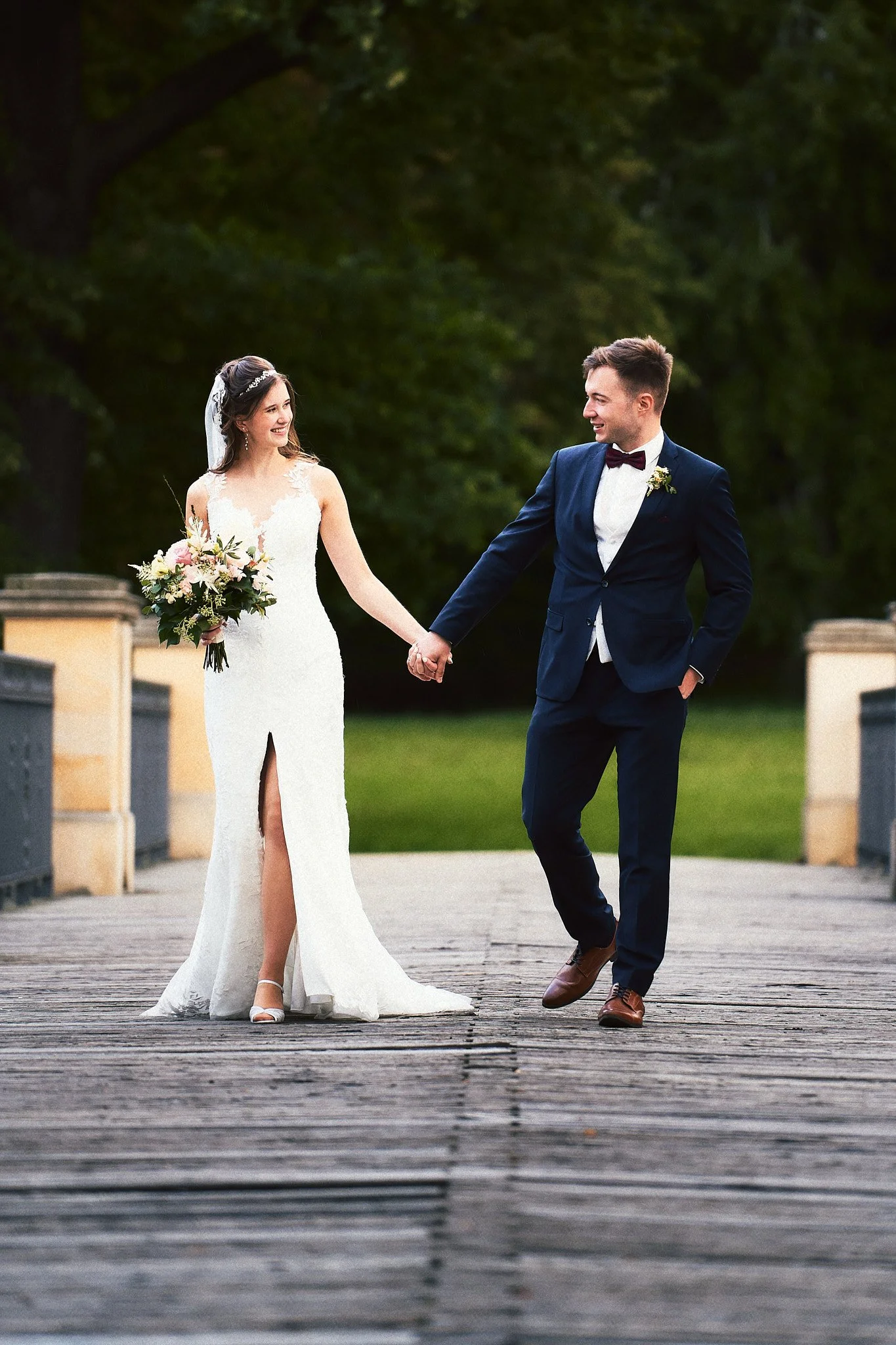 Ein Brautpaar hält Händchen beim Spazieren auf einem Holzsteg im Freien, umgeben von Bäumen, bei Sonnenuntergang.