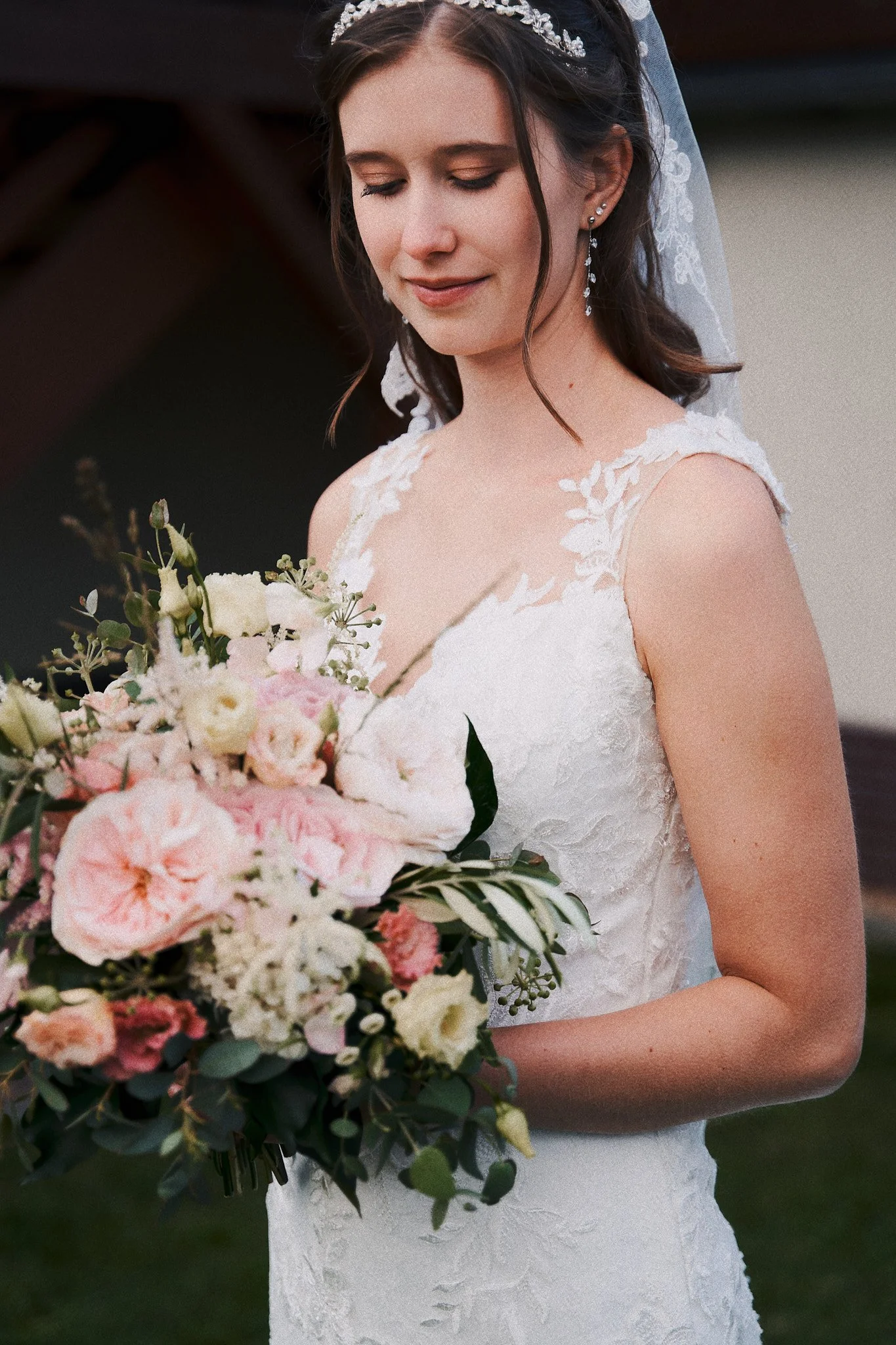 Eine junge Braut trägt ein elegantes weißes Hochzeitskleid mit floraler Spitze, hält einen großen Blumenstrauß aus rosa und weißen Blumen, trägt lange Ohrringe, ein Haarband mit Perlen und ein durchsichtiges Schleierkopfschmuck, während sie lächelnd 