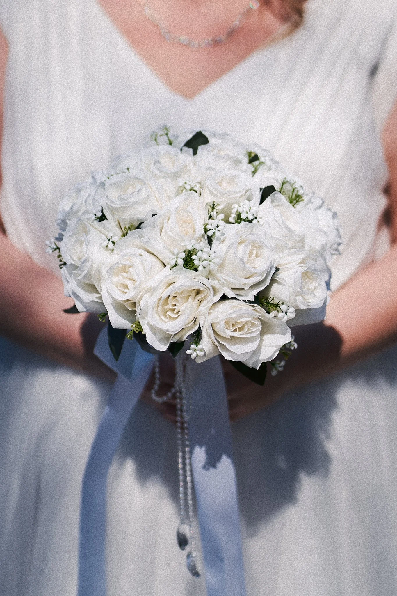 Frau in weißem Brautkleid hält einen weißen Brautstrauß mit Rosen und kleinen weißen Blüten.