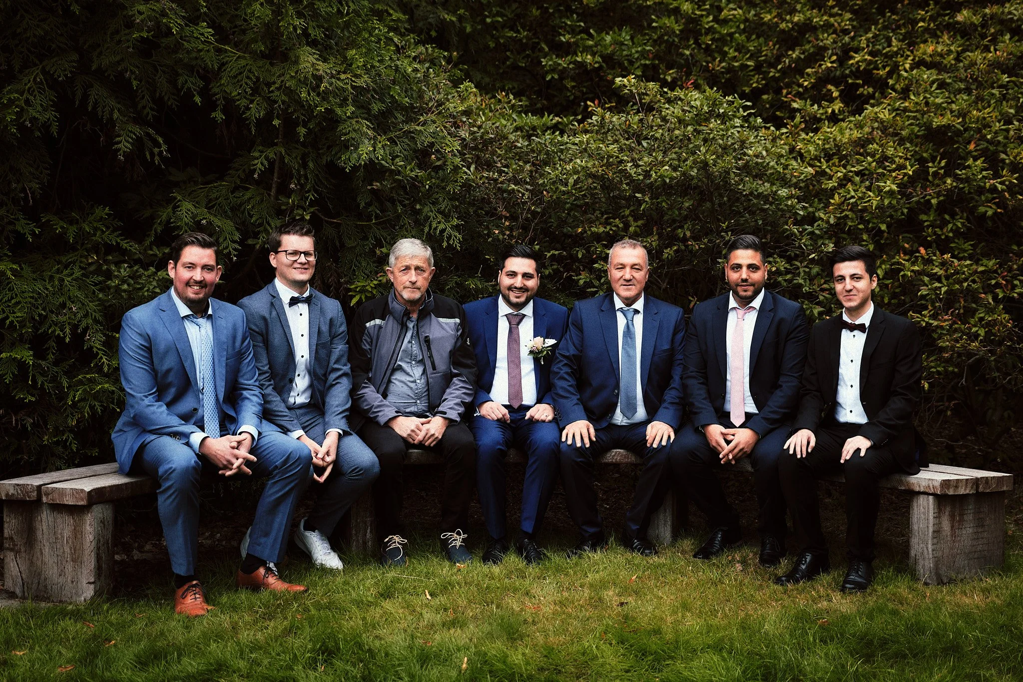Sieben Männer in formeller Kleidung sitzen auf einer Holzbank im Grünen, einige tragen Anzüge und Krawatten, einer trägt eine Jacke. Hinter ihnen dichtes Grün. Es sieht aus wie eine Hochzeits- oder Familienaufnahme.