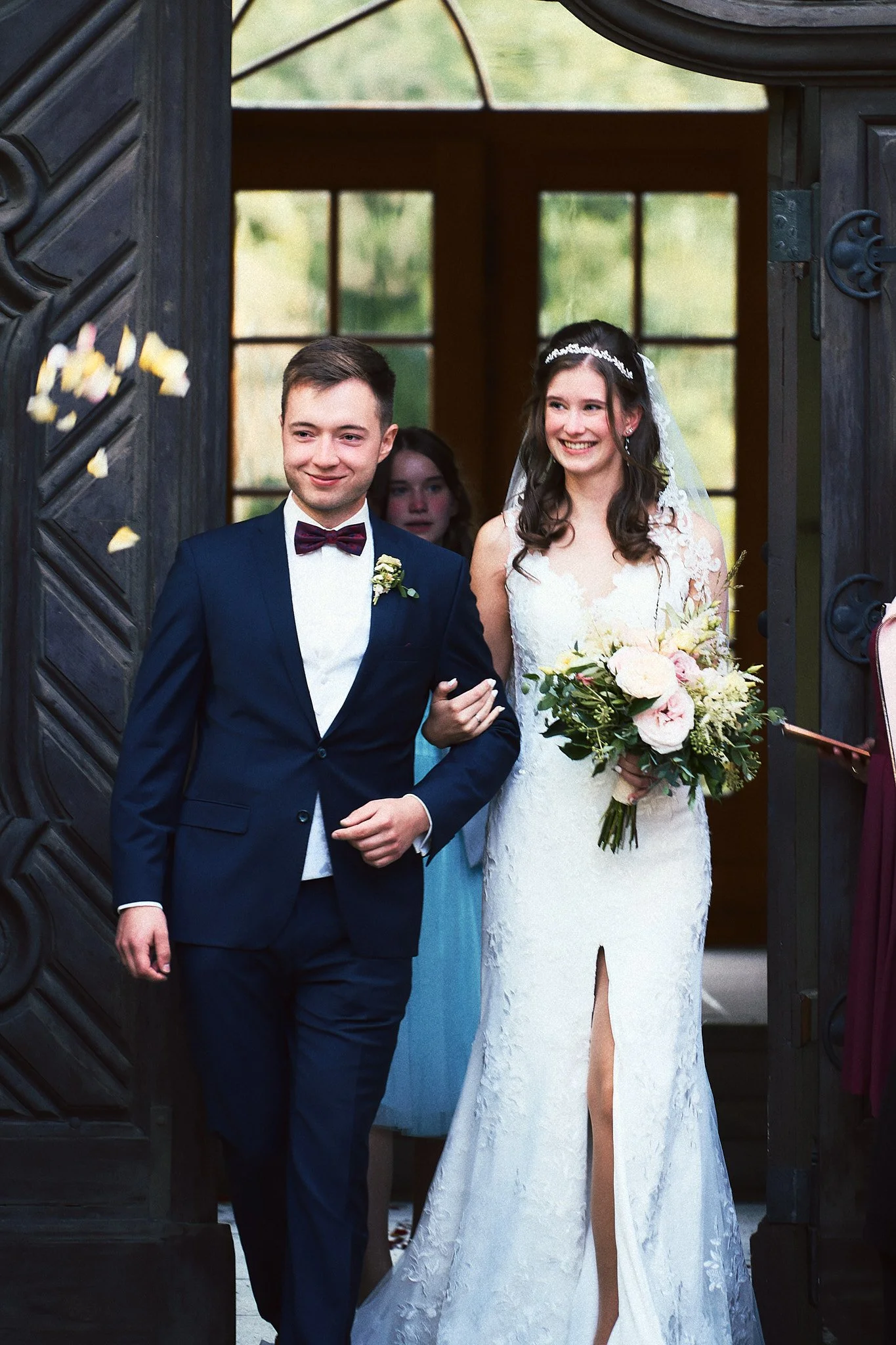 Braut und Bräutigam beim Hochzeitsauszug, lachend und in festlicher Kleidung, mit Blumenstrauß, beim Ausgehen aus einem dunklen Holzgebäude.