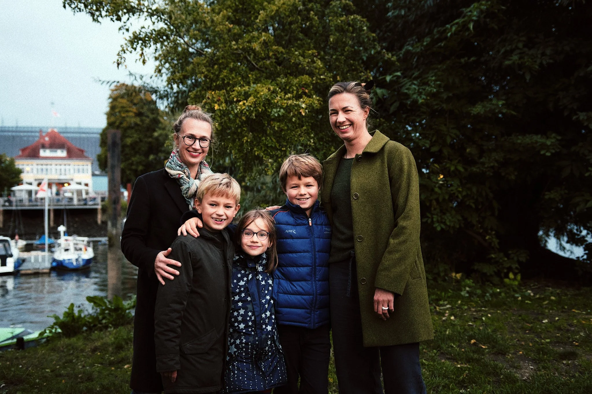 Eine Gruppe von fünf Personen, drei Kinder und zwei Erwachsene, die im Freien an einem Fluss mit Booten im Hintergrund lachen und posieren.
