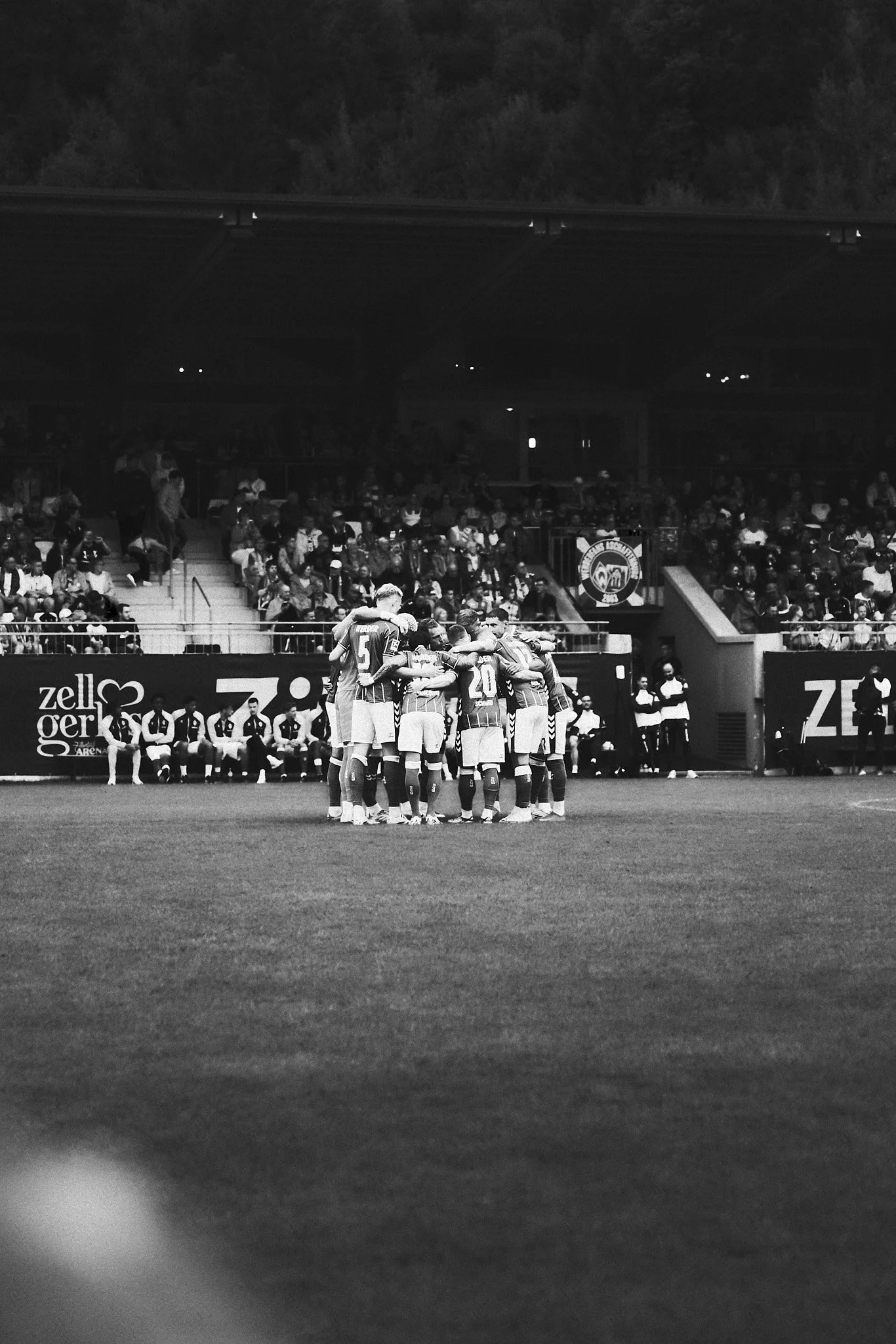 Schwarz-weiß-Foto von einem Fußballspiel, bei dem eine Mannschaft sich im Kreis versammelt und eingehend berührt, während die Sitzplätze voll mit Zuschauern sind, im Hintergrund sind Werbetafeln zu sehen.