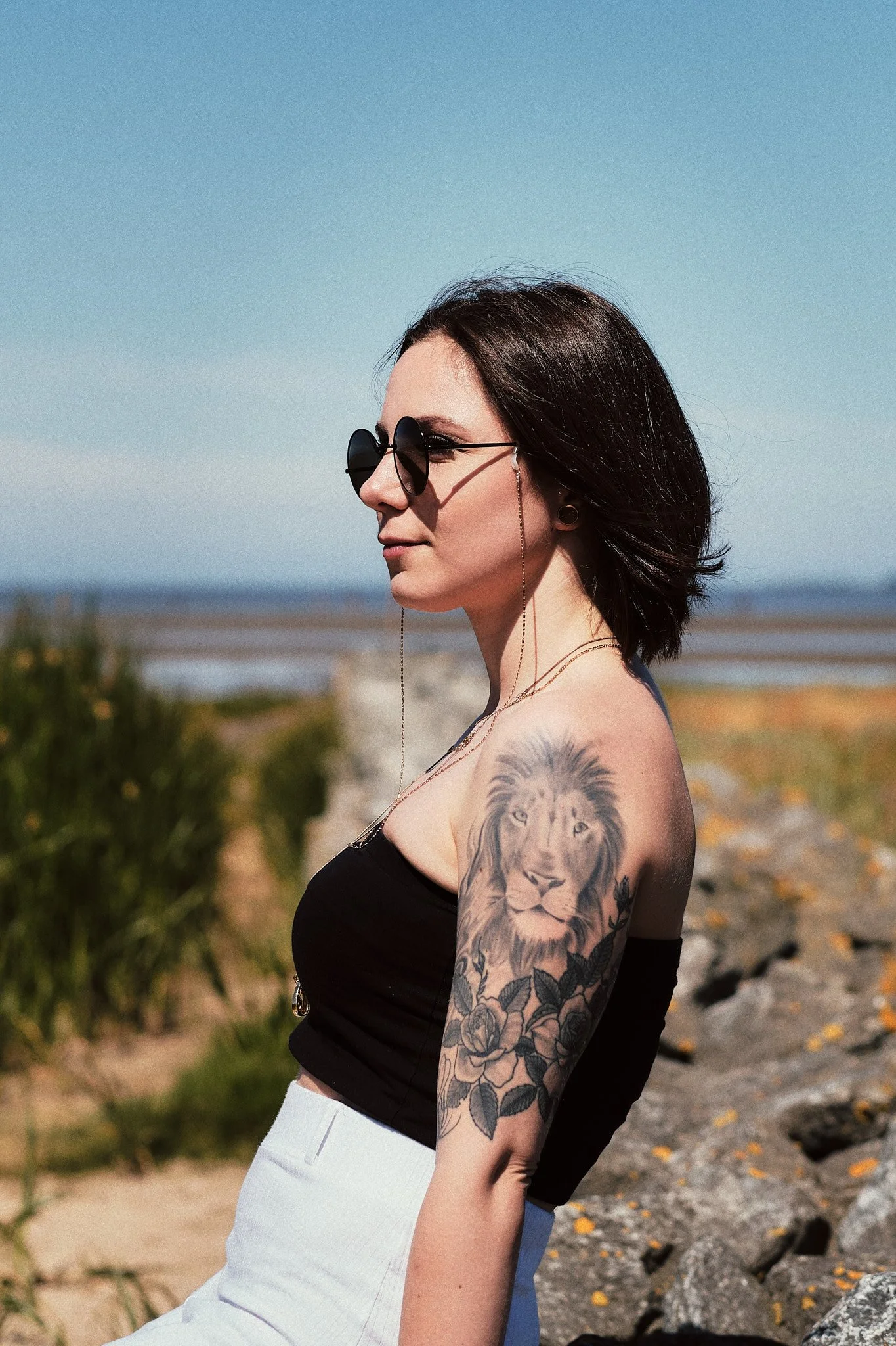Eine junge Frau mit dunklen Haaren, Sonnenbrille und Tattoo eines Löwen auf dem Oberarm steht im Freien bei sonnigem Wetter, mit einer Meereslandschaft im Hintergrund.
