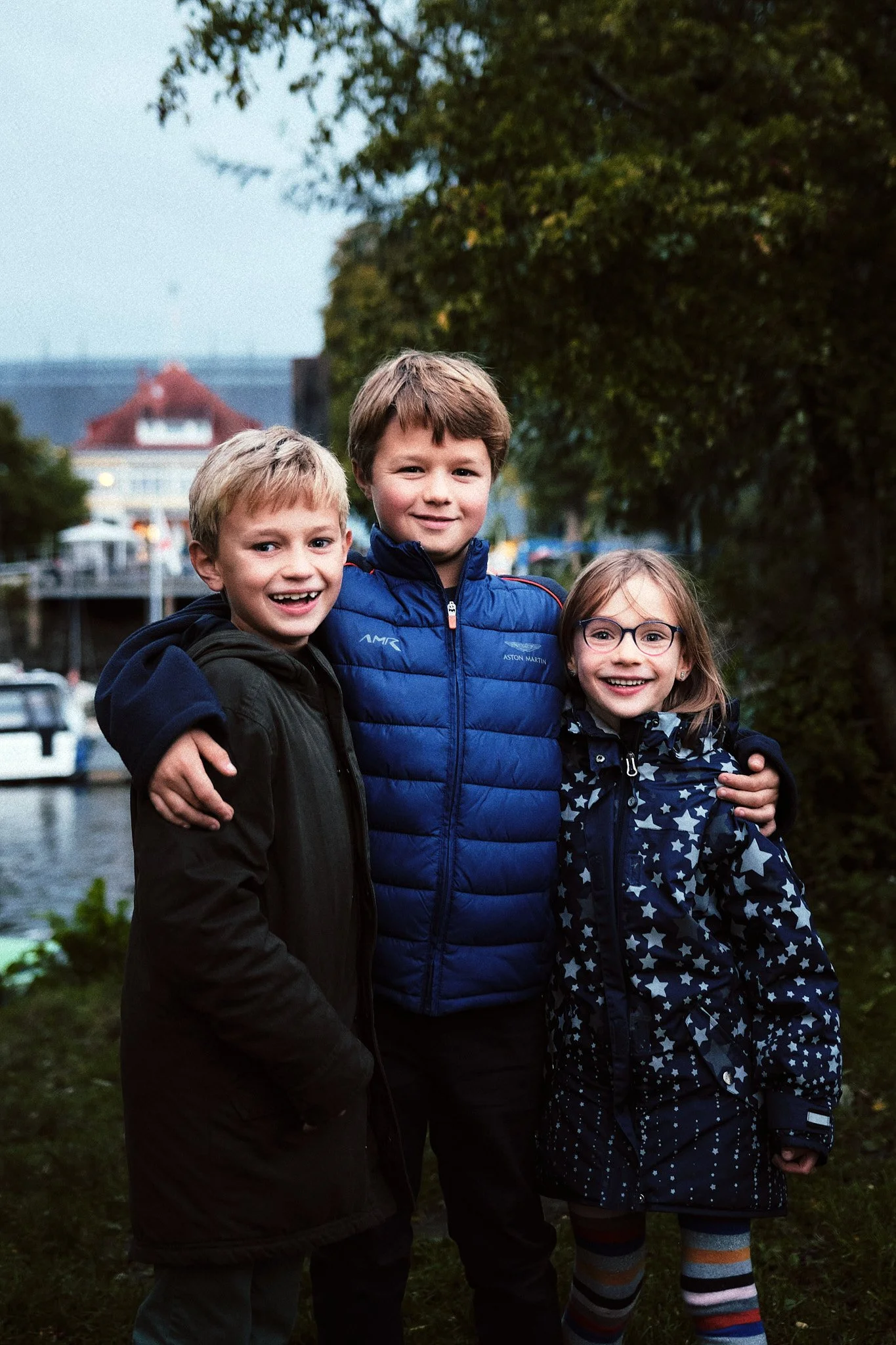 Drei lachende Kinder stehen draußen nahe am Wasser, umgeben von Bäumen, an einem bewölkten Tag.