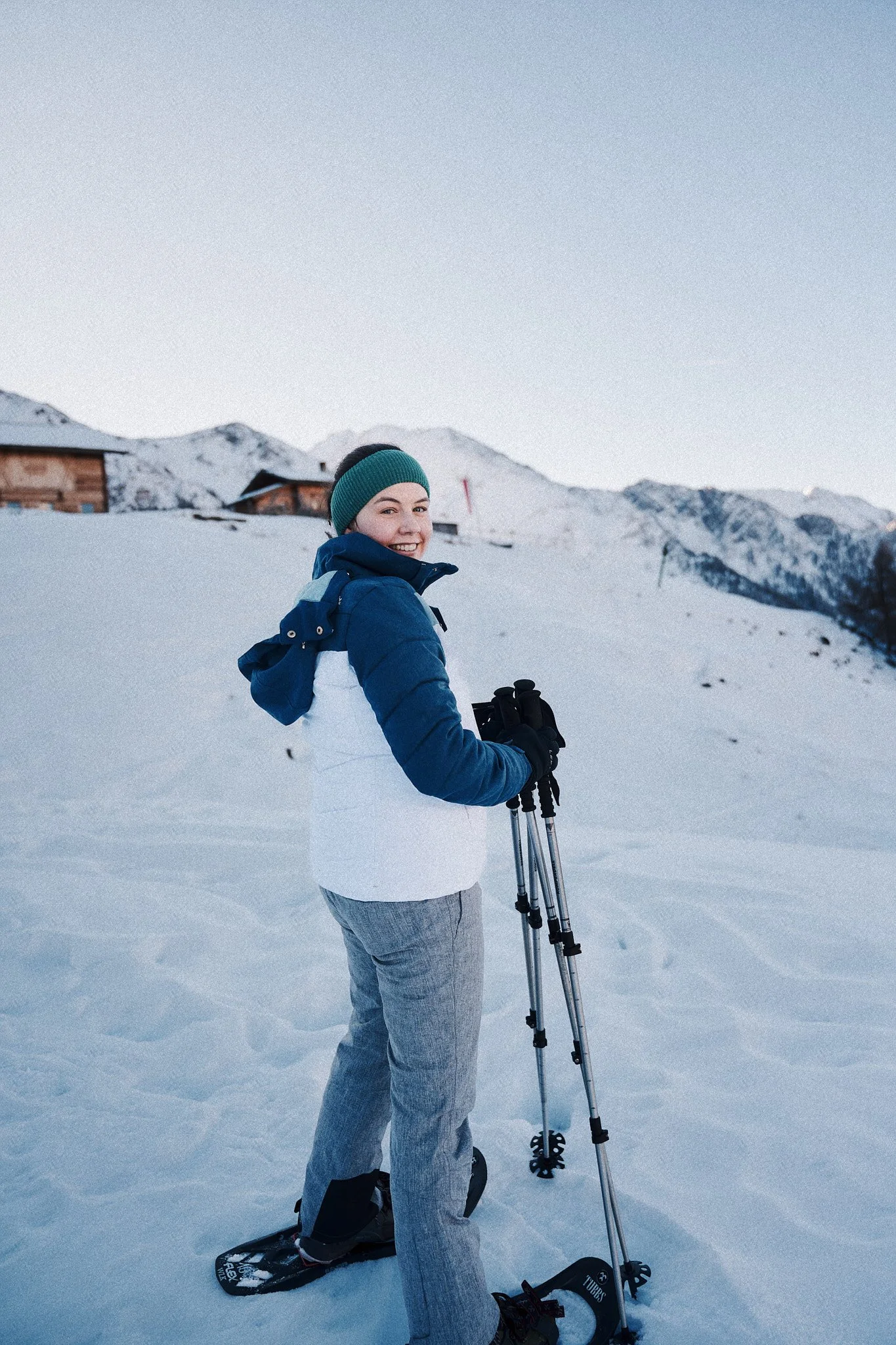 Eine Frau in Winterkleidung steht mit Skiern und Skistöcken im Schnee in den Bergen, hinter ihr sind Hütte und Berge sichtbar.