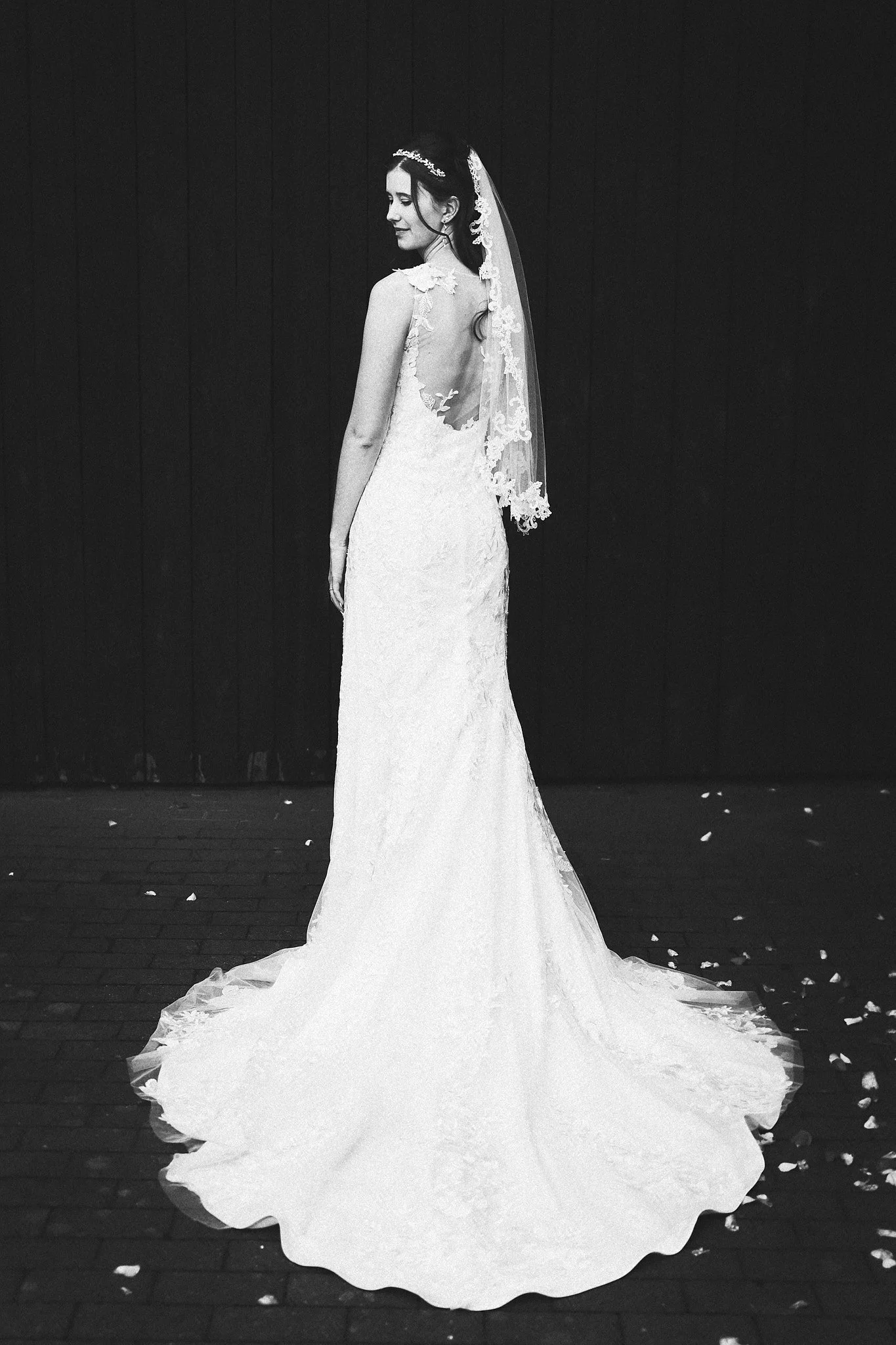 Eine Braut trägt ein langes, elegantes weißes Hochzeitskleid mit einem offenen Rücken und Spitzenverzierungen, sie steht vor einem dunklen Hintergrund, und verstreute Blumenblätter liegen am Boden.