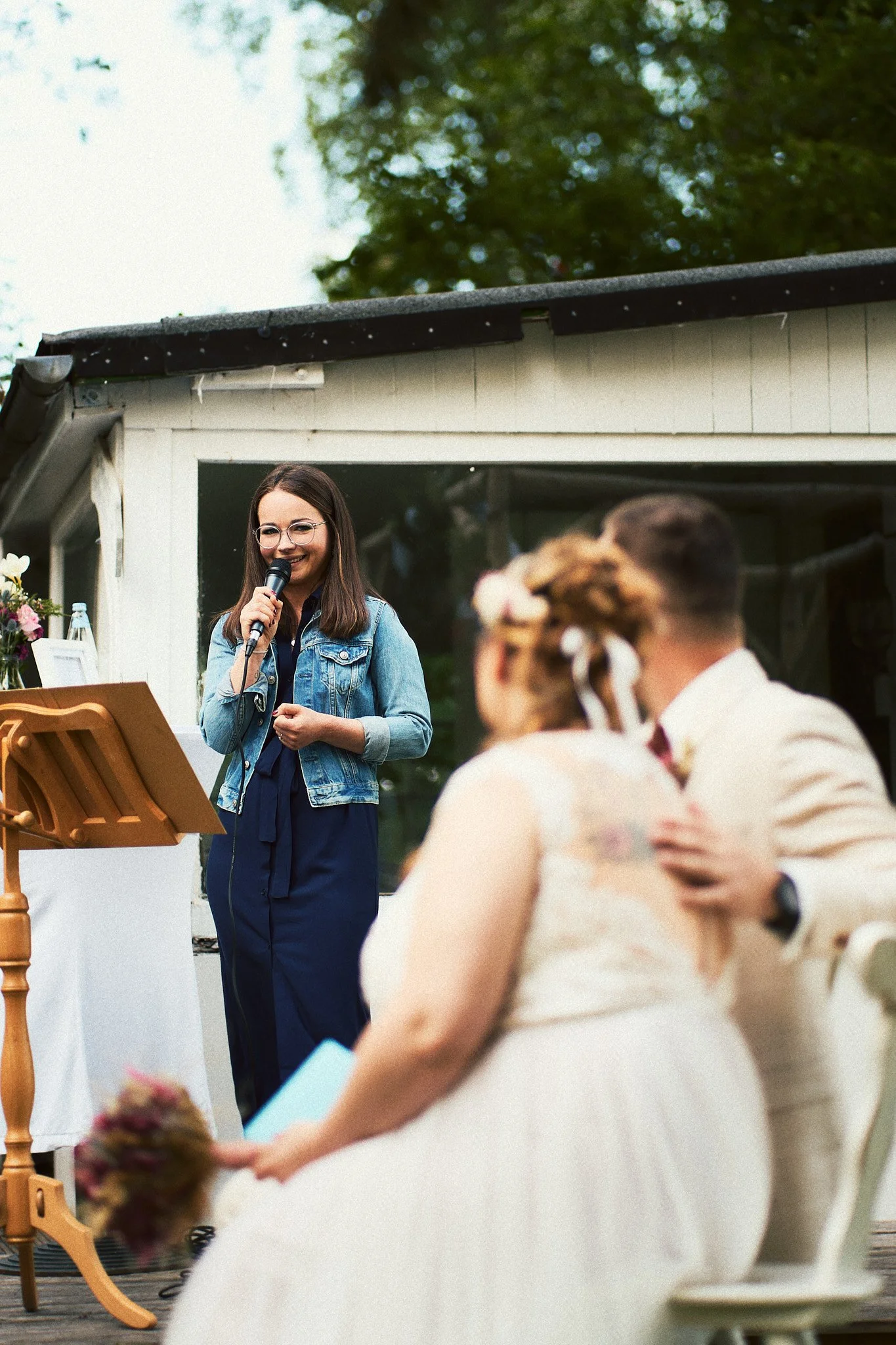 Eine junge Frau hält eine Mikrofon und spricht bei einer Outdoor-Hochzeit, im Vordergrund sind Braut und Bräutigam unscharf abgebildet, die auf einen Sitzplatz vor einem Holzhaus sitzen.