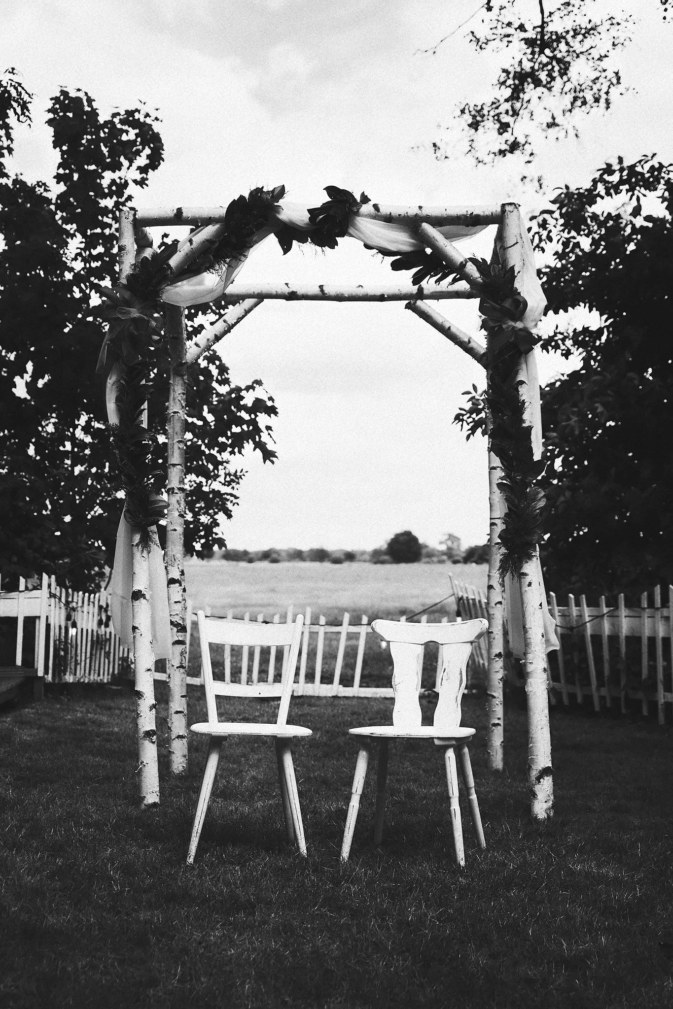 Zwei weiße Holzstühle vor einem Bogengemach mit grünen Blättern, im Freien, auf einer Wiese, in Schwarzweiß.