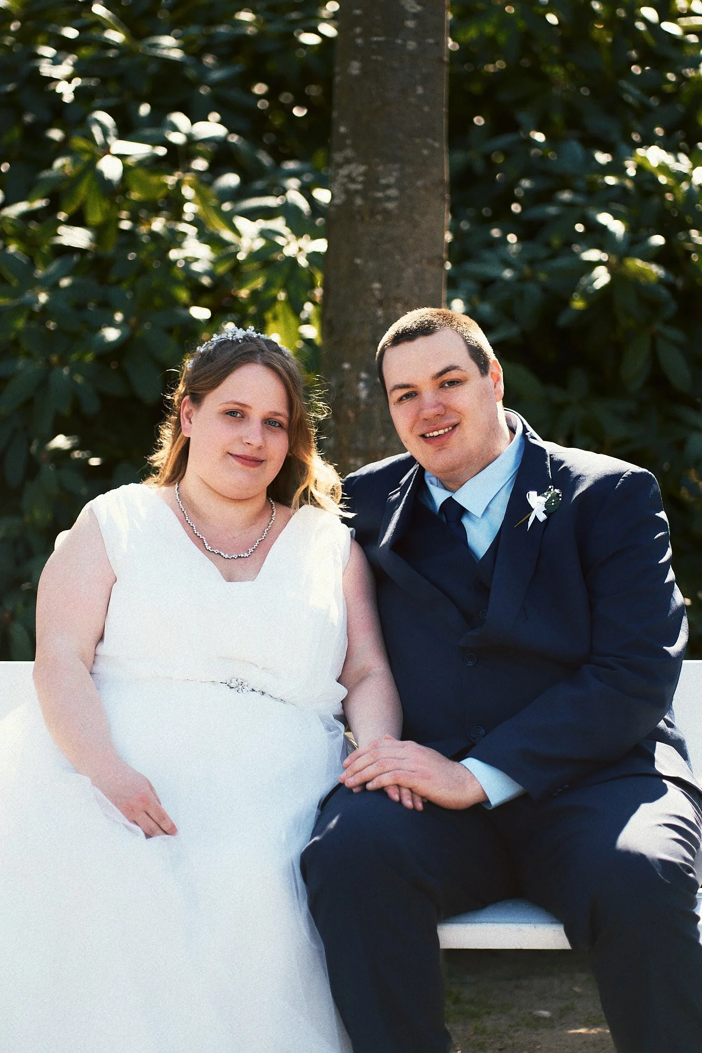 Ein Brautpaar sitzt auf einer Bank im Garten, die Braut trägt ein weißes Hochzeitskleid und eine Perlenkette, der Bräutigam trägt einen dunklen Anzug mit Krawatte.