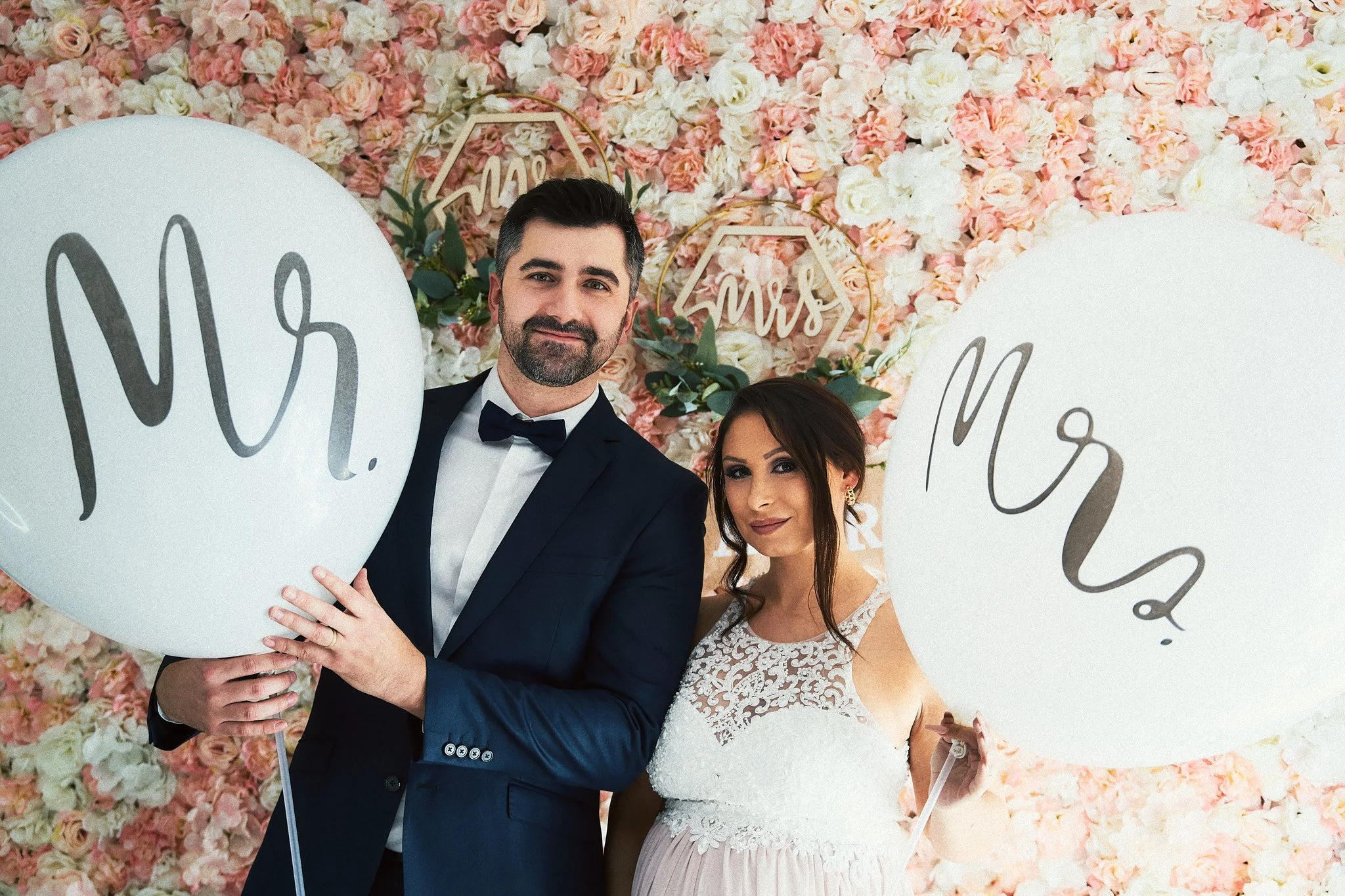 Ein Brautpaar bei einer Hochzeit, hält große weiße Ballons mit der Aufschrift 'Mr.' und 'Mrs.' vor einem Hintergrund aus rosa und weißen Blumen.