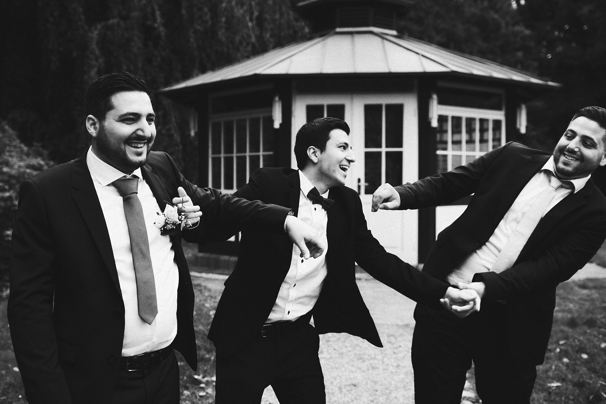 Drei Männer in Anzügen lachen und spielen zusammen vor einem kleinen Gartenhaus im Freien.