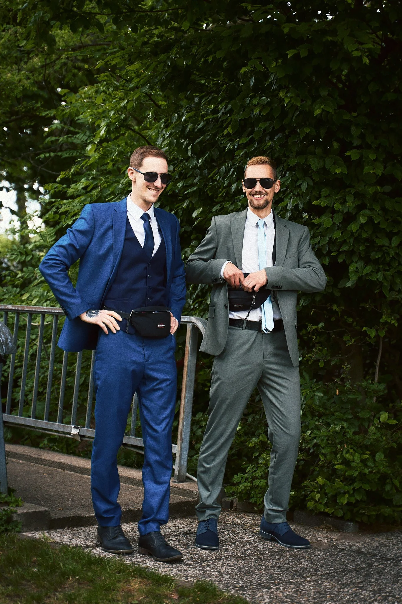 Zwei Männer in Anzügen und Sonnenbrillen stehen vor einem grünen Busch und lachen.