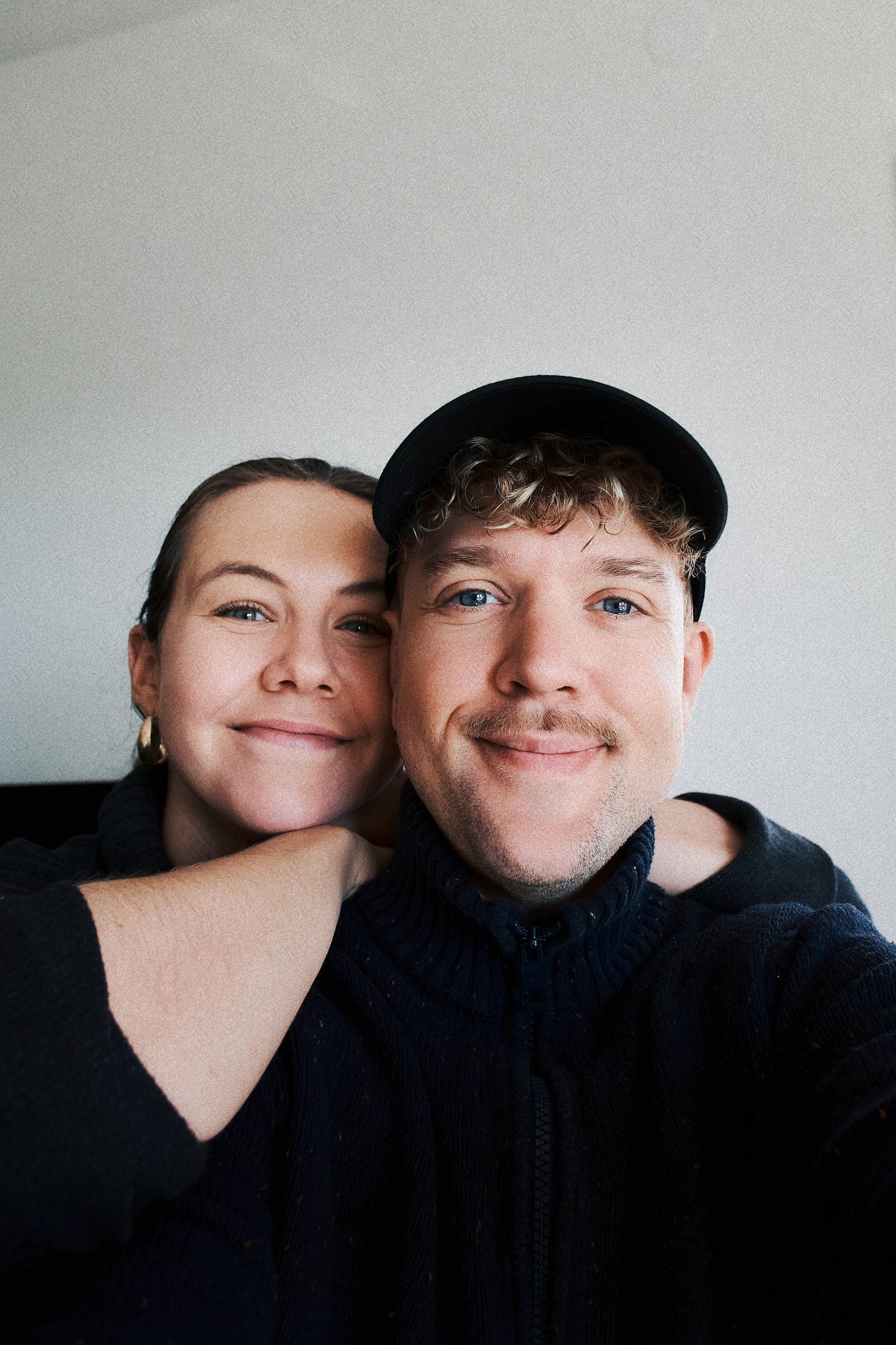 Ein lächelndes Paar macht ein Selfie, der Mann trägt einen schwarzen Pullover und eine schwarze Mütze, die Frau hat braune Haare und trägt goldene Ohrringe.