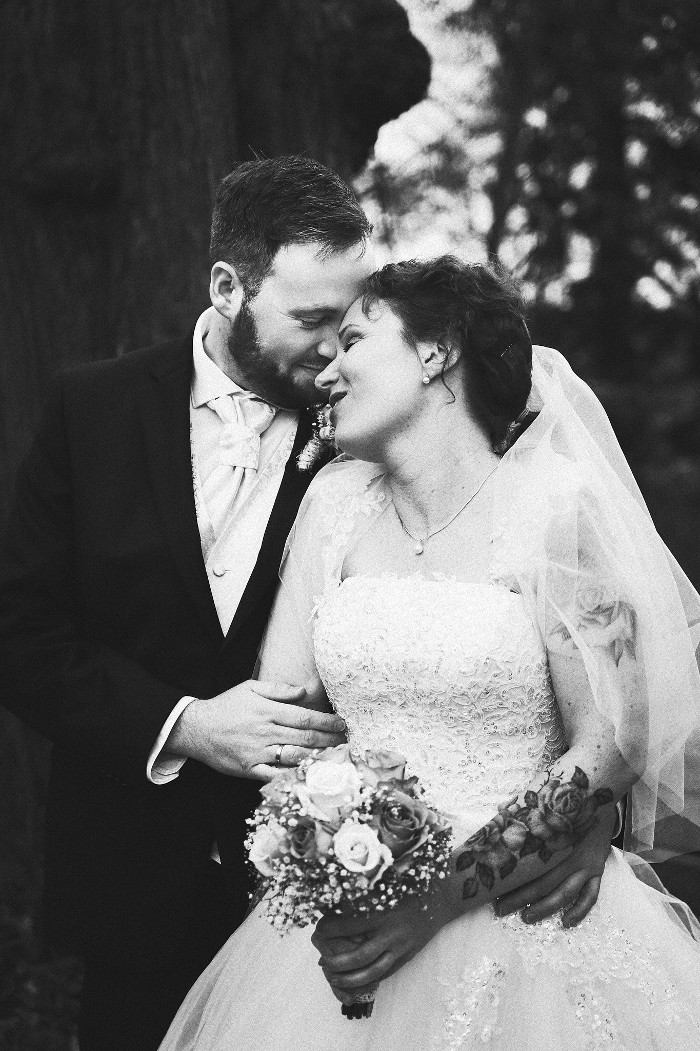 Ein Brautpaar bei einer Hochzeit, beide umarmen sich liebevoll, im Hintergrund Bäume, in Schwarzweiß.