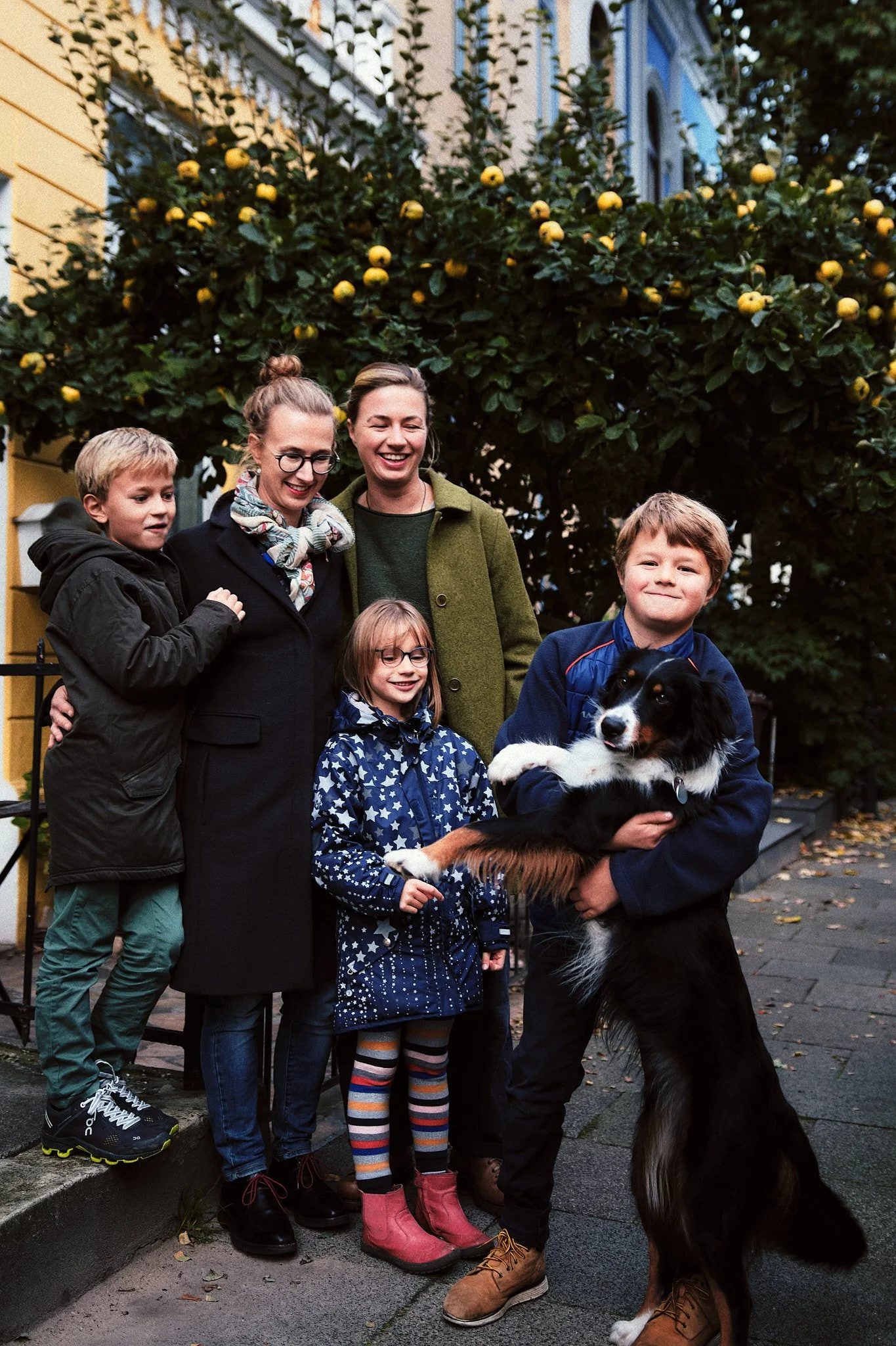 Familie mit Kindern und Hund auf der Straße, im Hintergrund ein Baum mit gelben Früchten.