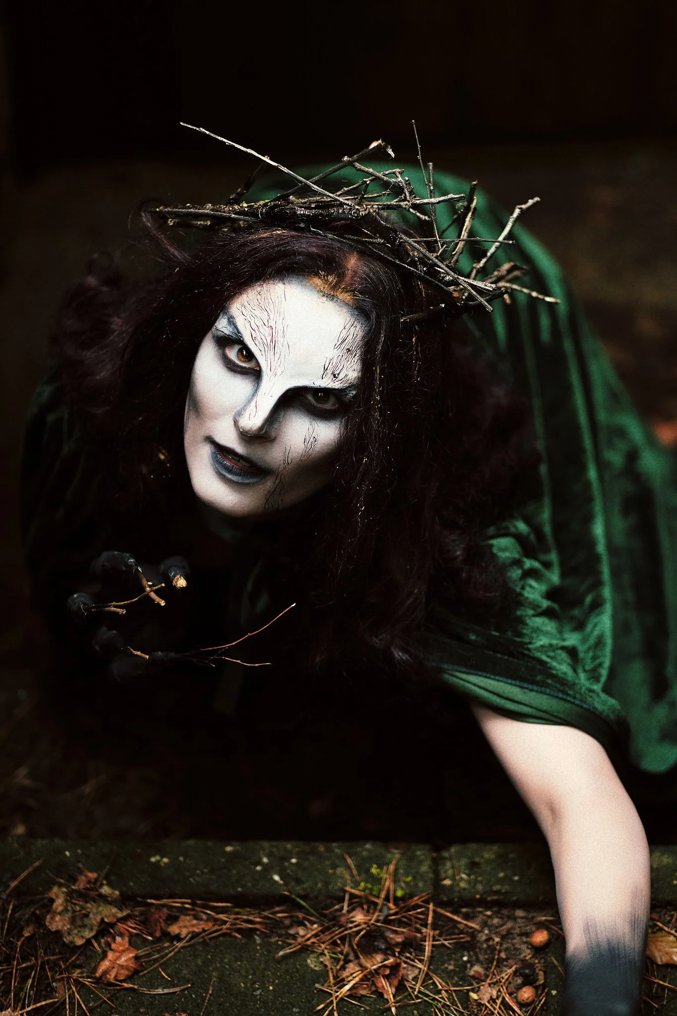 Person in dunklem Grün mit schwarzem Haar, weiße Gesichtsbemalung, trägt einen Kranz aus Ästen auf dem Kopf, liegt auf dem Boden im Freien.