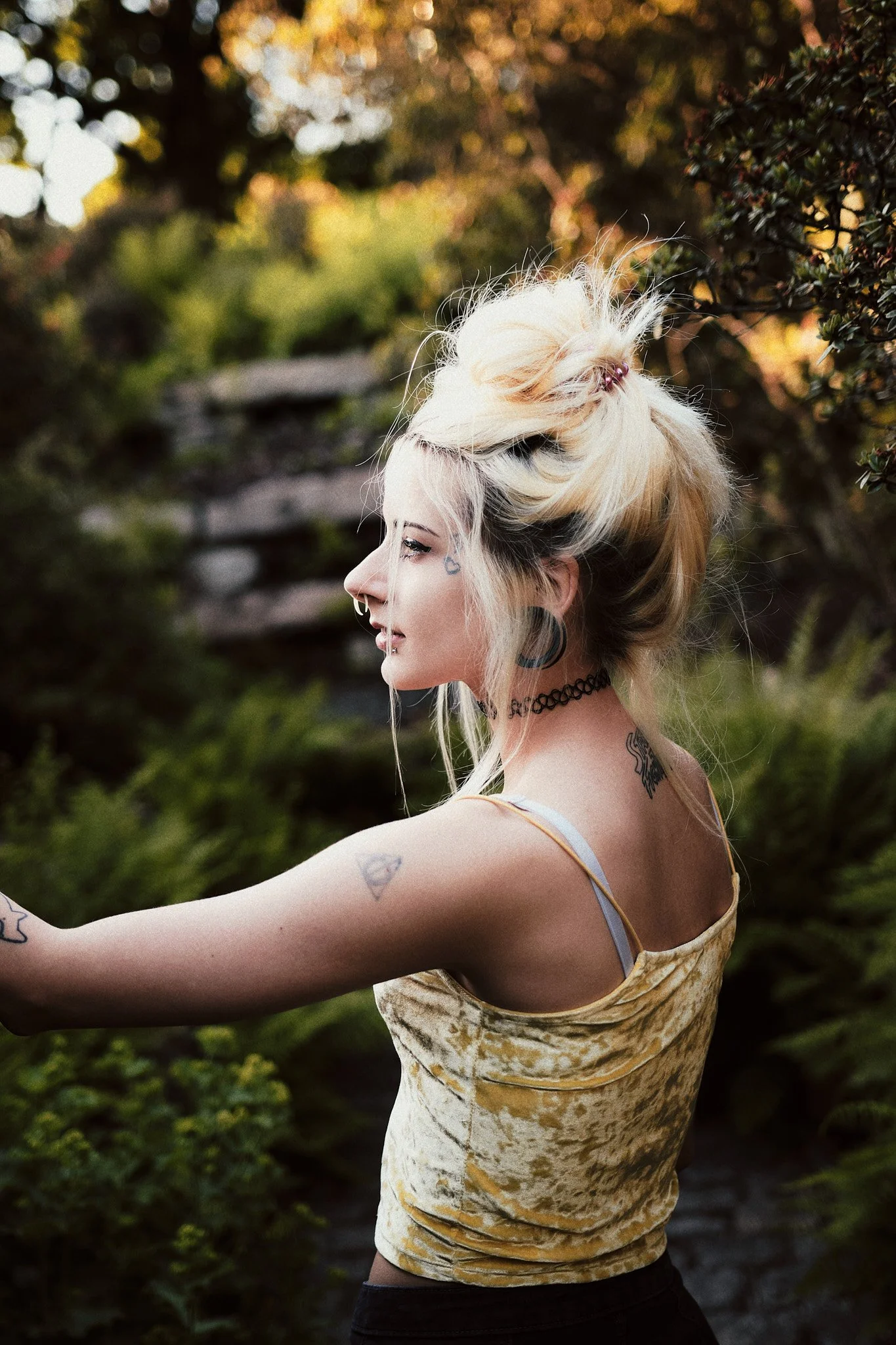 Frau mit Tattoos und blonder Haare im Grünen, bei Sonnenuntergang, seitlich betrachtet.