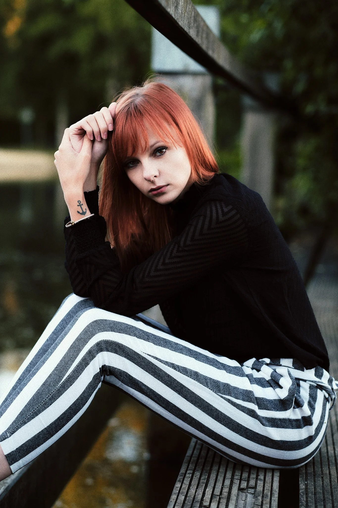 Junge Frau mit roten Haaren, schwarz gekleidet, sitzt auf einer Bank an einem Fluss, Blick in die Kamera, Tattoo am Handgelenk, auf der Jacke sind Zickzack-Muster, im Hintergrund Bäume und Wasserspiegel.