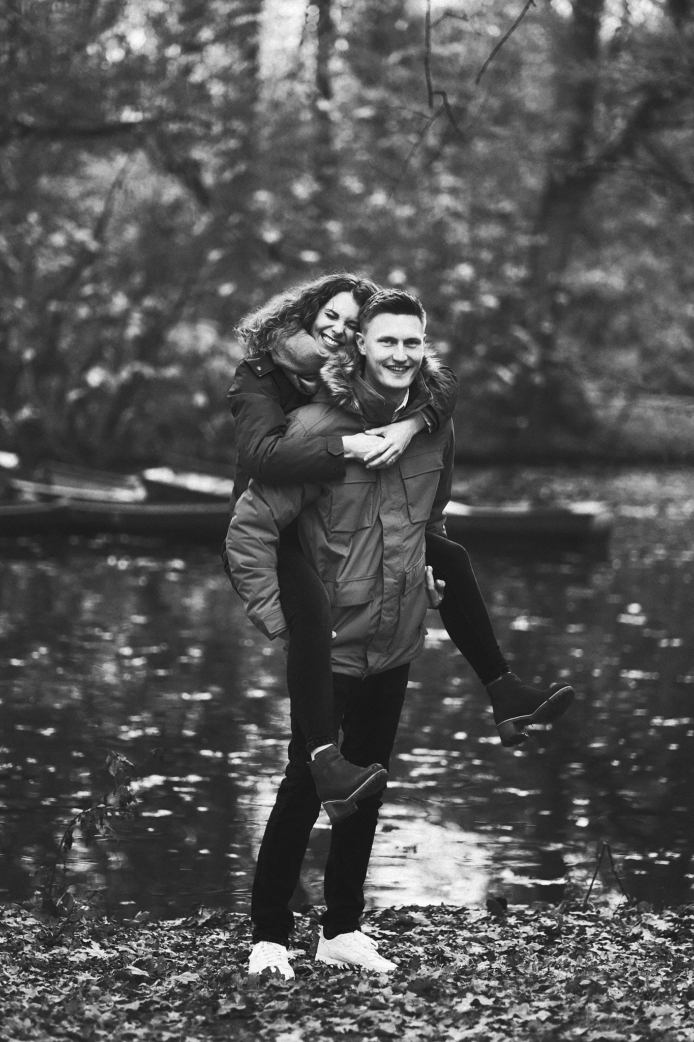 Ein Paar in einem Wald am Flussufer, der Mann trägt eine Jacke und trägt die Frau auf den Schultern, beide lachen.