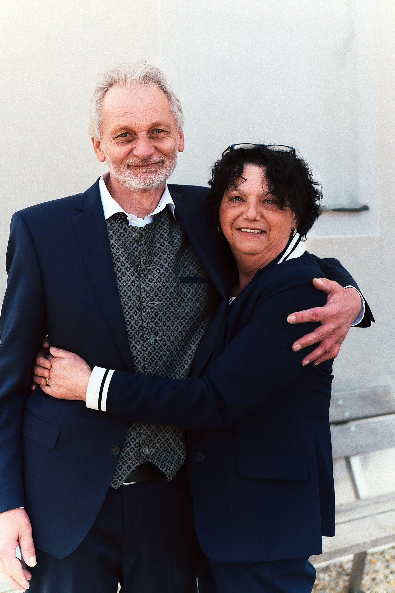 Zwei wohlbekleidete Personen umarmen sich lachend vor einer hellen Wand