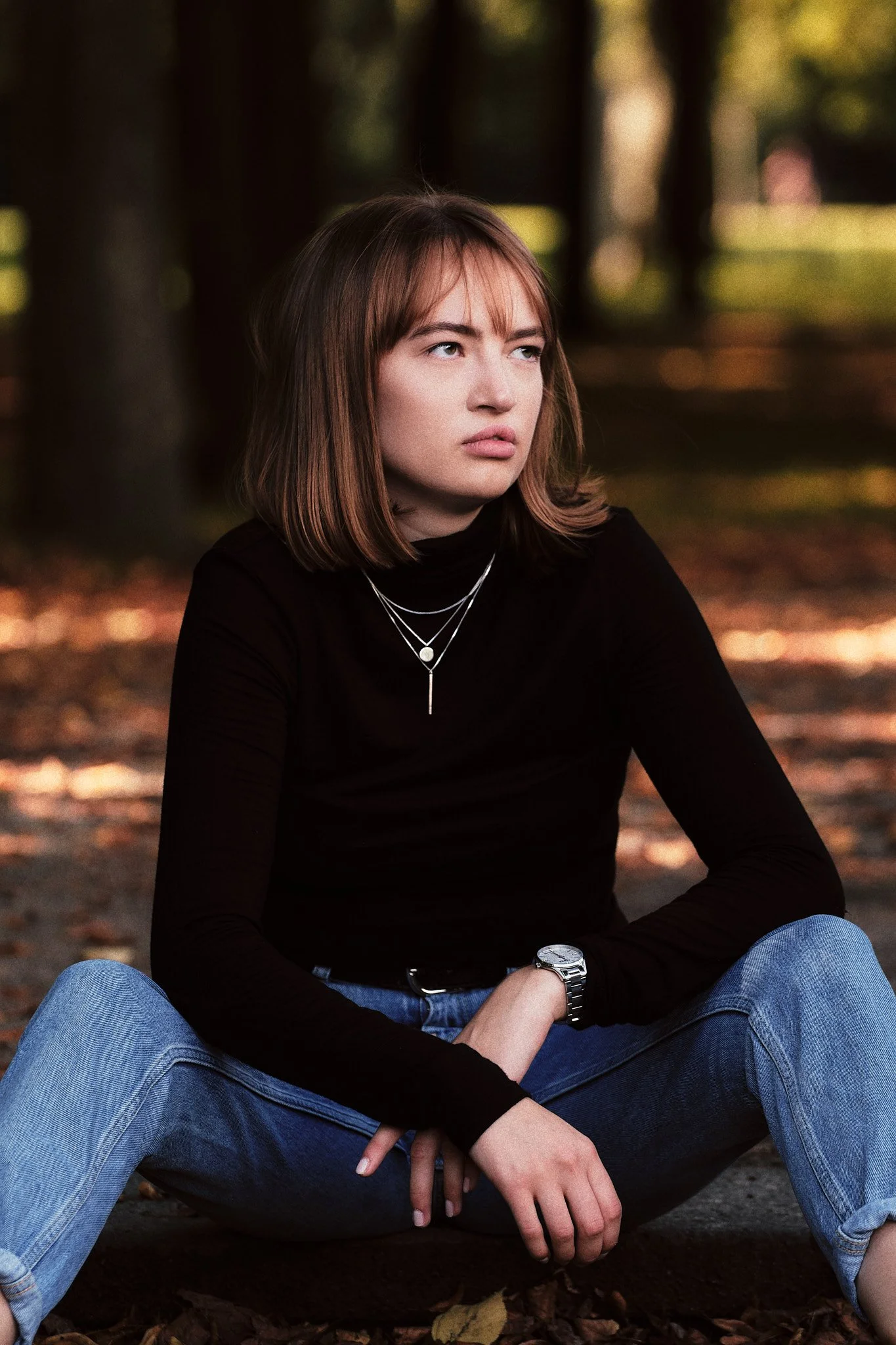 Junge Frau sitzt auf dem Boden im Wald, trägt schwarzes T-Shirt, blaue Jeans, silberne Ketten und Armband, hat braune Haare, schaut nachdenklich.