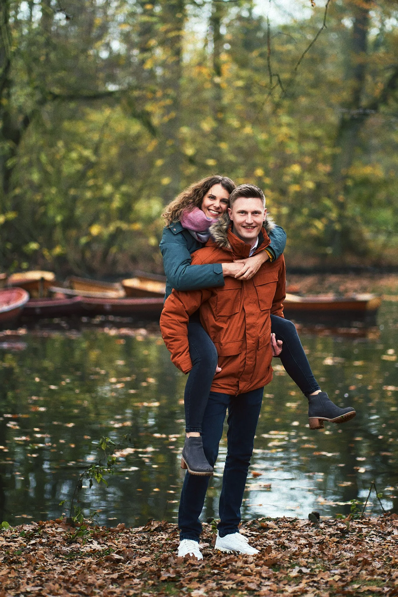 Ein Paar bei einem Herbstspaziergang am See, der Mann trägt einen braunen Mantel und trägt die Frau auf seinem Rücken, im Hintergrund Boote auf dem Wasser und bunte Herbstbäume.