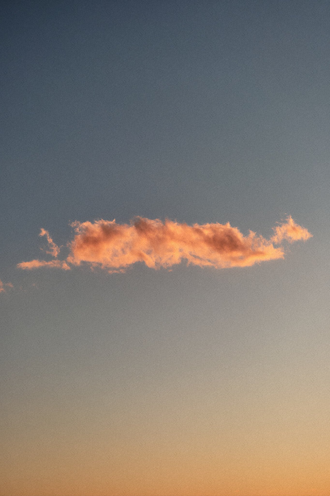 Ein orangefarbener Wolkenstreifen am Himmel bei Sonnenuntergang, mit einem Übergang von hellen bis dunklen Farbtönen.
