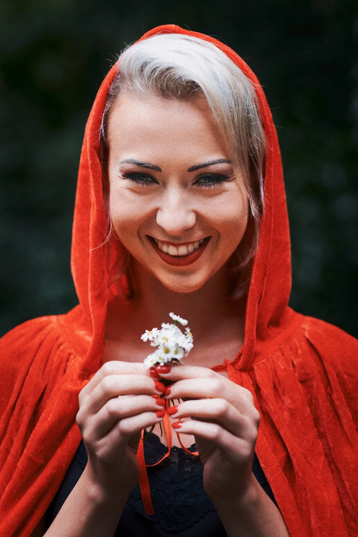 Junge Frau mit blonder, kurz geschnittener Frisur, trägt eine rote Kapuze, lächelt und hält kleine weiße Blumen in den Händen.