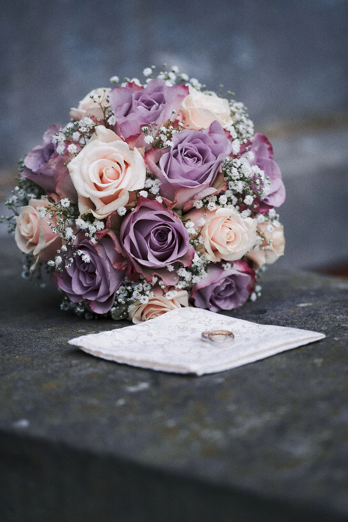 Ein Brautstrauß aus pastellfarbenen Rosen in Lilaverläufen und weißen Blumen, mit einem weißen Handtuch und einem Ehering.