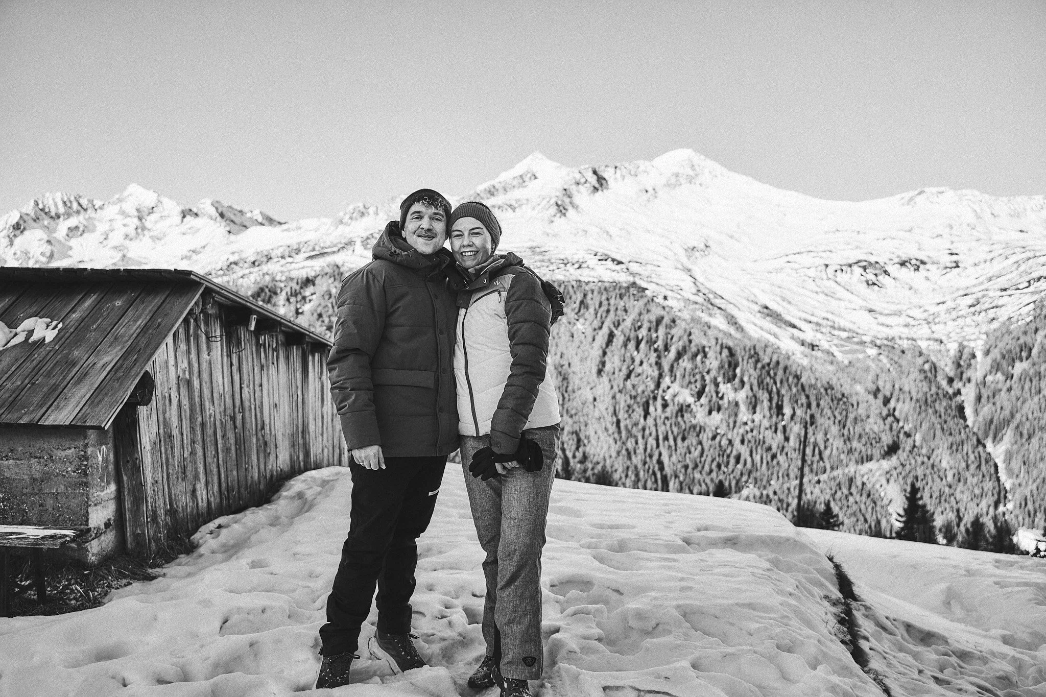 Ein glückliches Paar in Winterkleidung steht in Schneelandschaft vor einer Hütte mit verschneiten Bergen im Hintergrund.