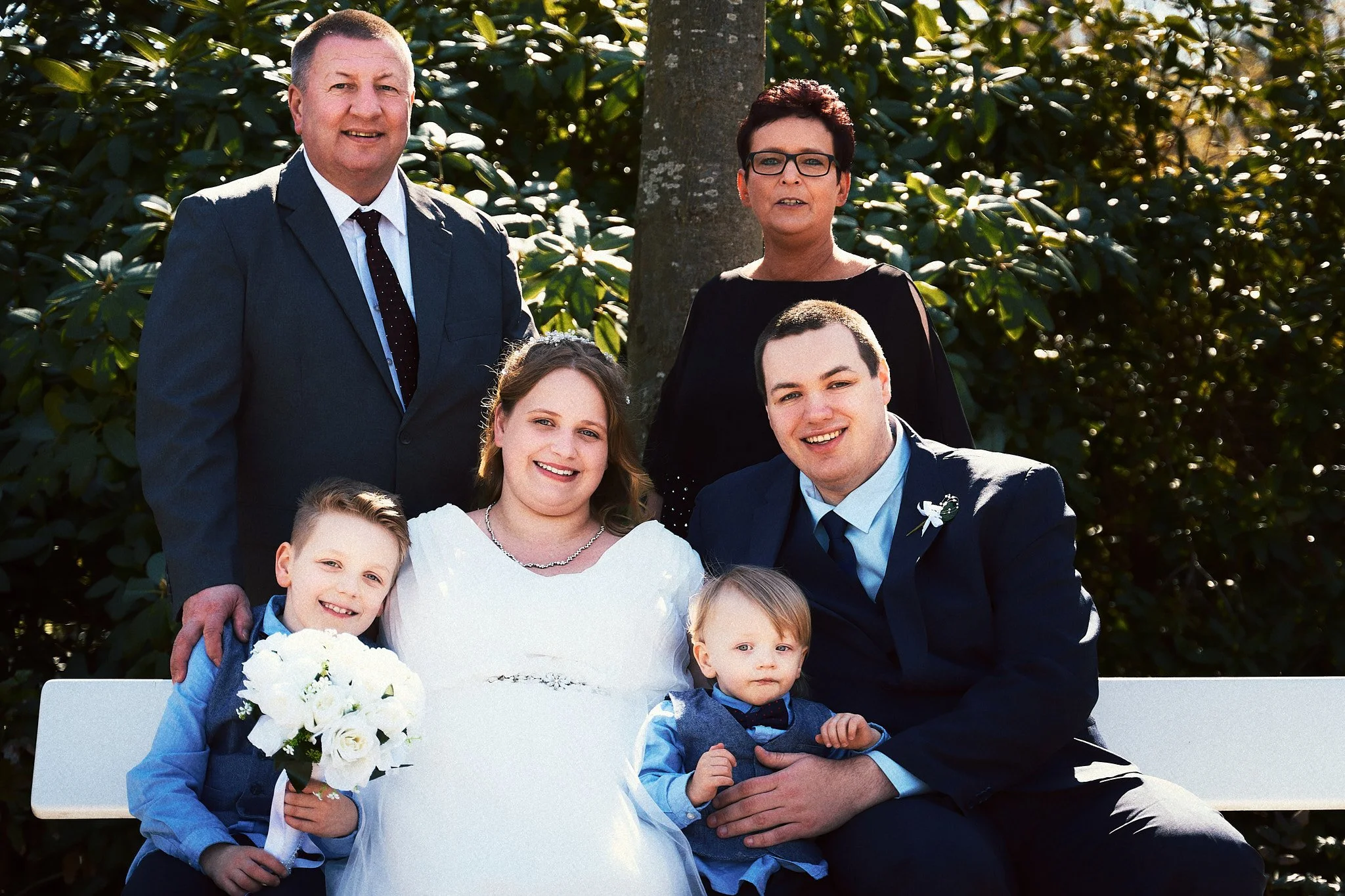 Familie bei einer Hochzeit im Freien, sitzend auf einer Bank, mit grünen Büschen im Hintergrund. Erwachsene, Kinder und ein Brautpaar sind zu sehen.