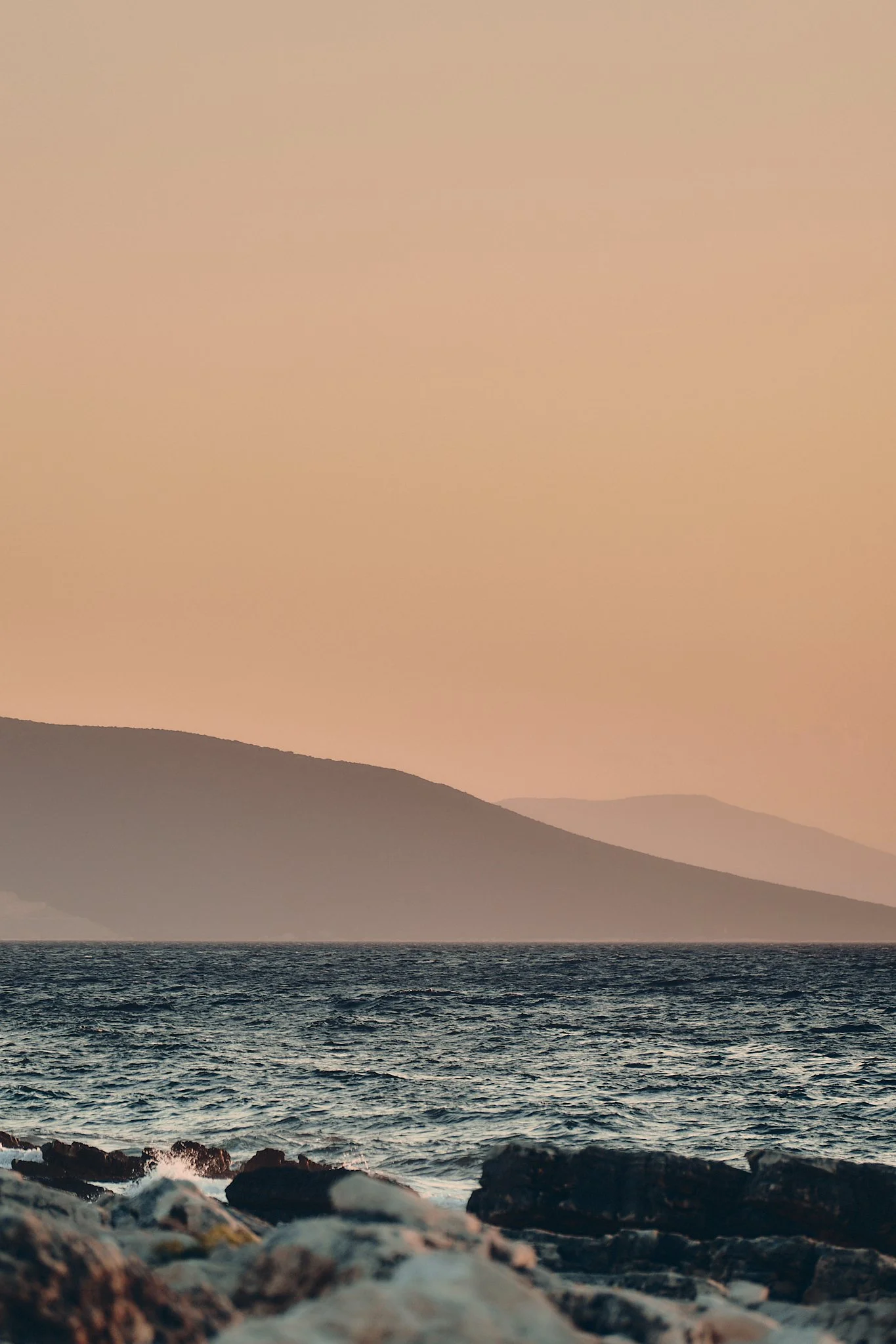 Blick auf das Meer mit Wellen, im Vordergrund Steine, im Hintergrund Hügel bei Sonnenuntergang.
