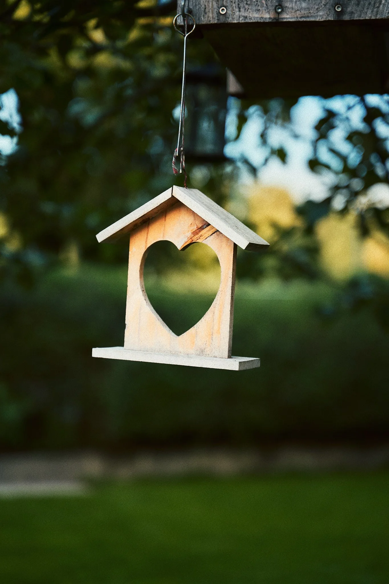 Ein hölzernes Vogelhaus mit einem Herz ausgeschnitten, hängt an einem Draht vor einem grünen, unscharfen Hintergrund.