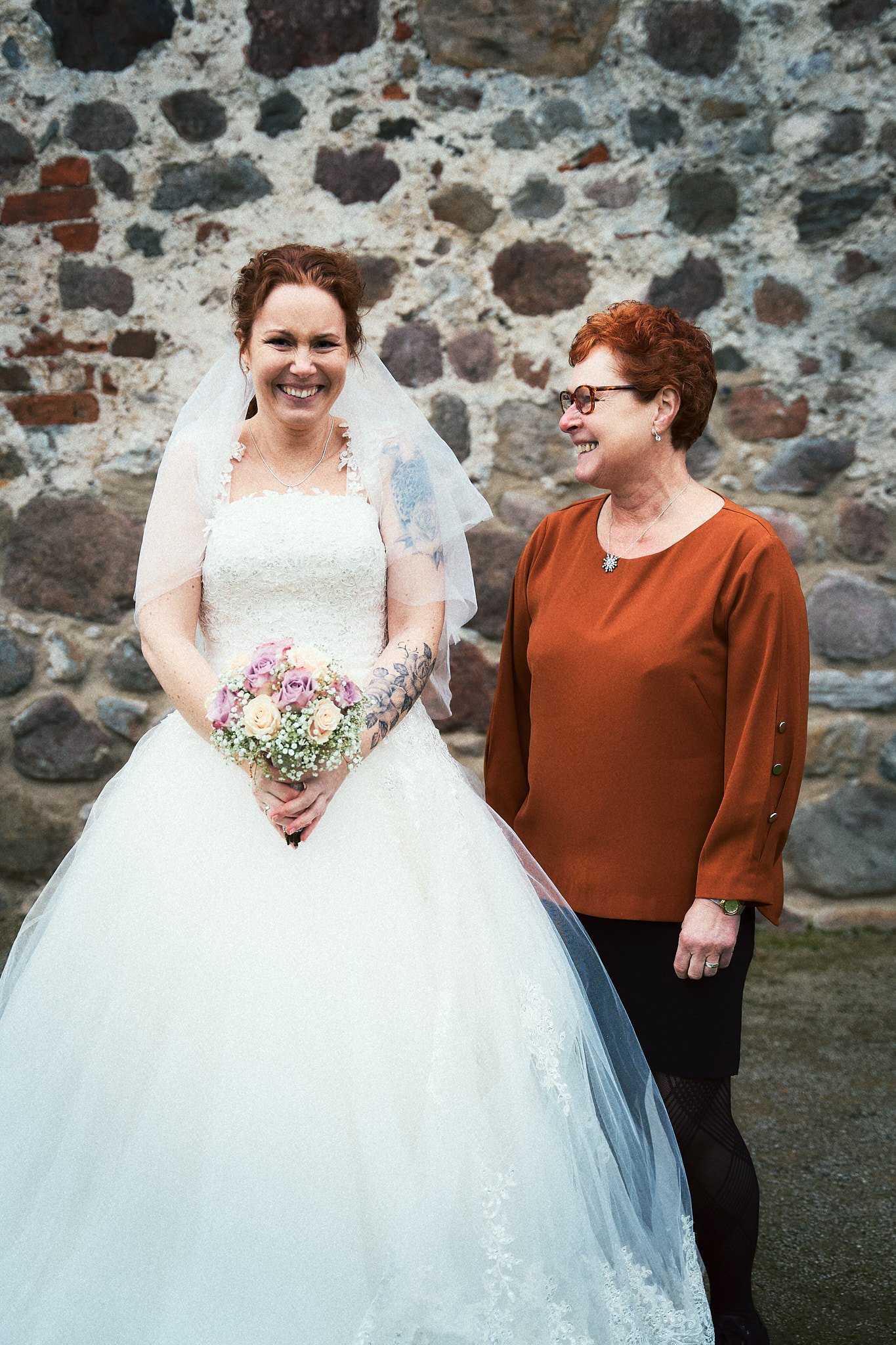 Eine Braut in weißem Kleid mit Blumenstrauß steht neben einer lächelnden Frau in braunem Oberteil vor einer Steinewand.