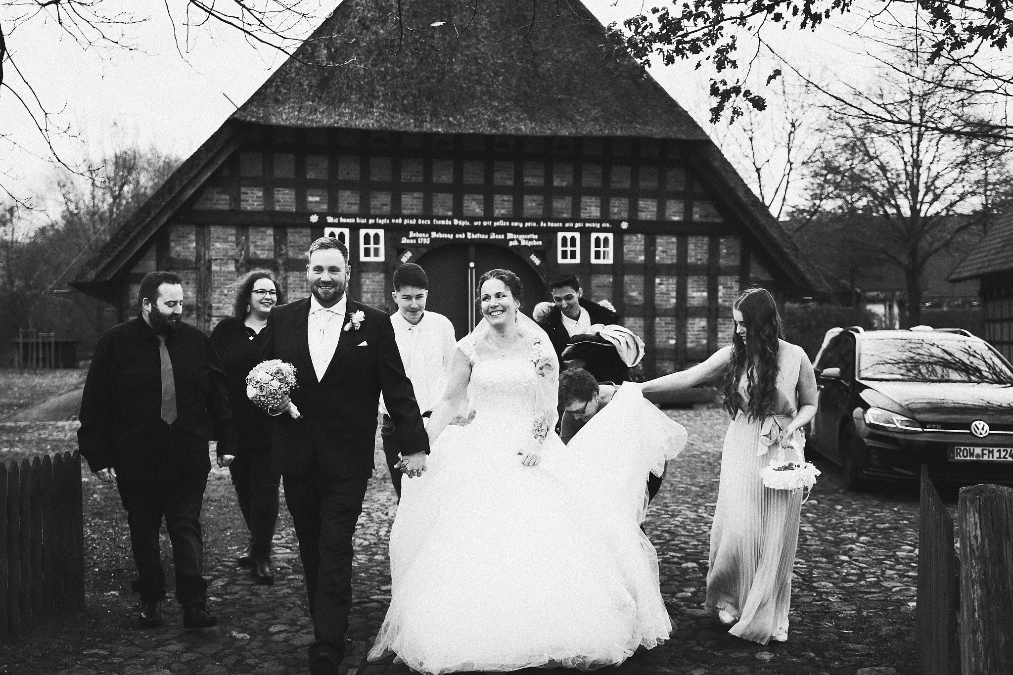 Eine Hochzeitsgesellschaft läuft gemeinsam vor einem alten Fachwerkhaus in schwarzer und weißer Fotografie, um einen besonderen Anlass zu feiern.
