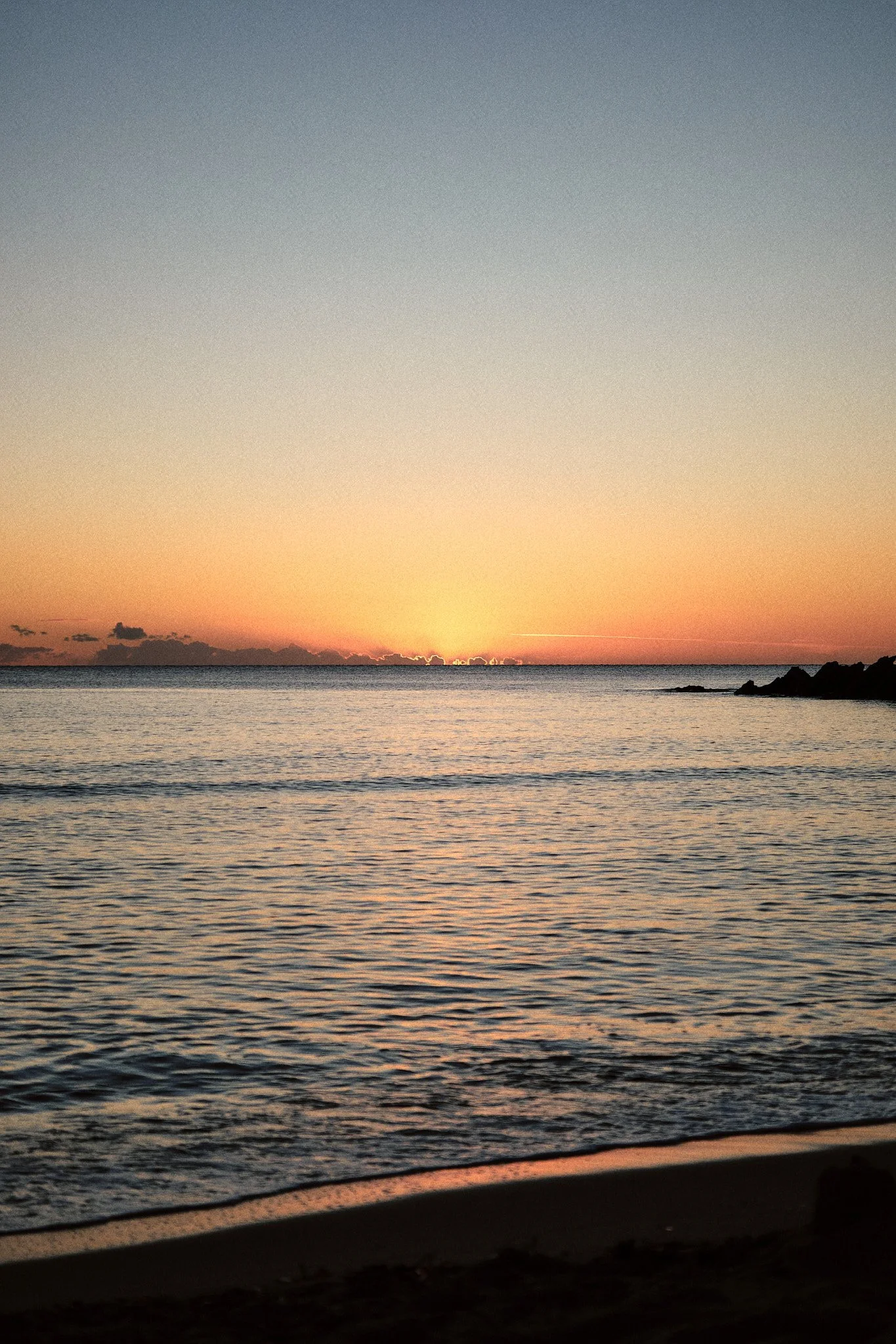 Ein Sonnenuntergang über dem Ozean mit einem klaren Himmel, ruhigem Wasser und einem dunklen Strand im Vordergrund.