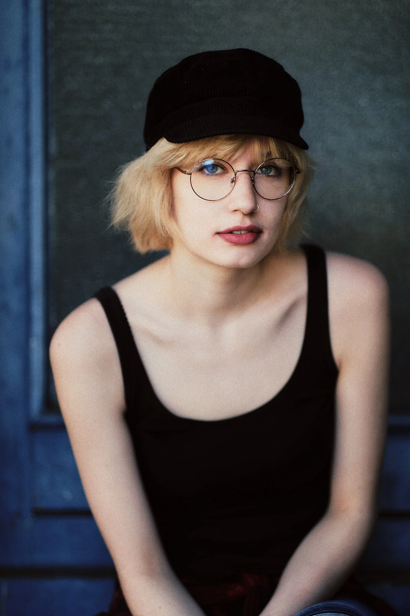 Junge Frau mit blondem, kurz gewelltem Haar, Brille, Nasenpiercing und schwarzen Kappe, trägt schwarzes Top, sitzt vor einer grauen Wand.