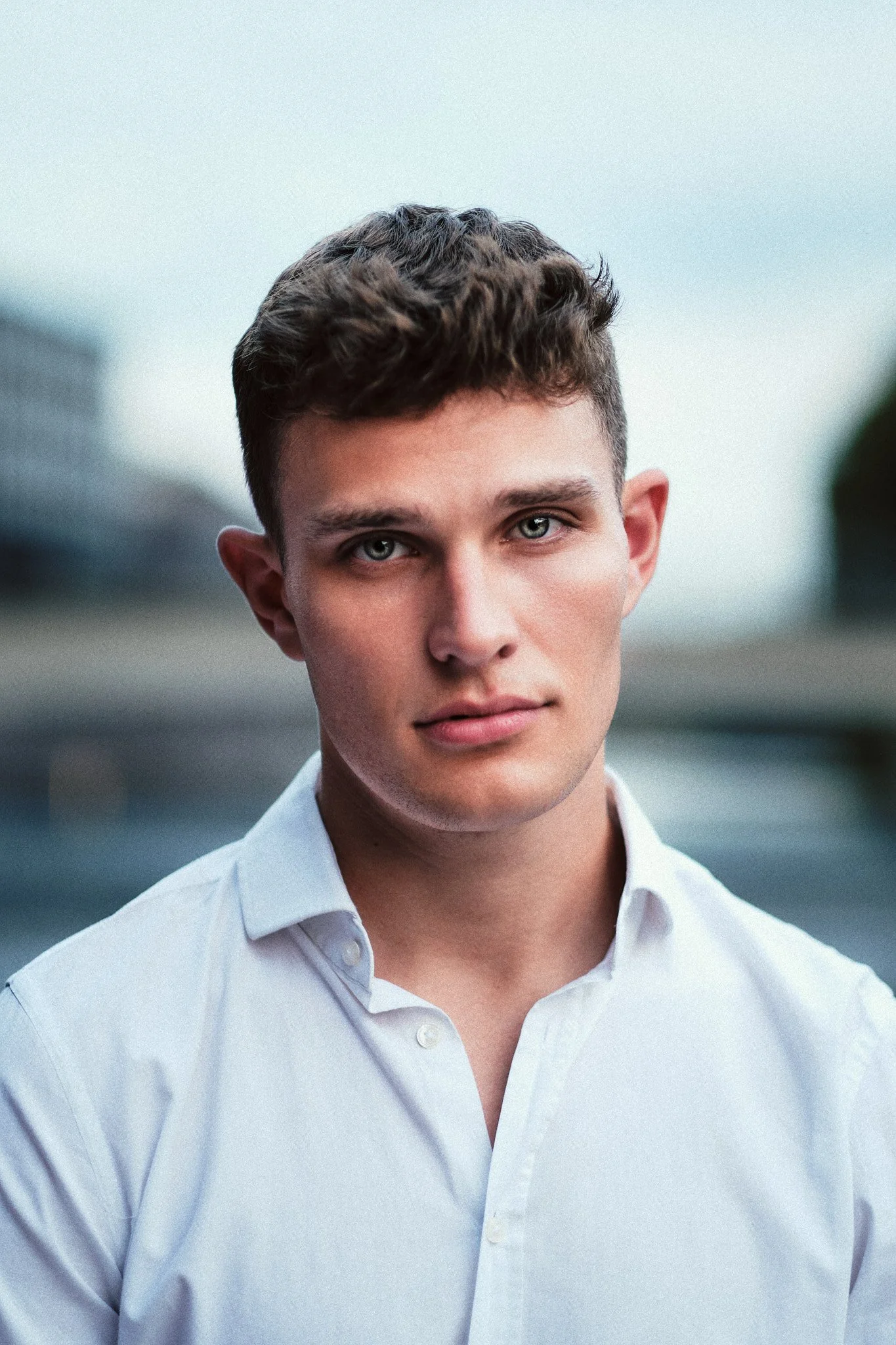 Nahaufnahme eines jungen Mannes mit kurzen braunen Haaren, blauen Augen und hellem Teint, trägt ein weißes Hemd im Freien, unsicher bei bewölktem Himmel.