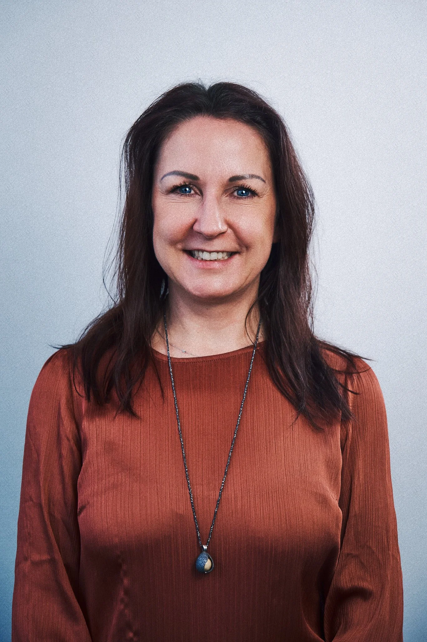 Portrait einer Frau mit dunkelbraunen Haaren, blauen Augen und einem braunen Oberteil, vor einem neutralen Hintergrund.