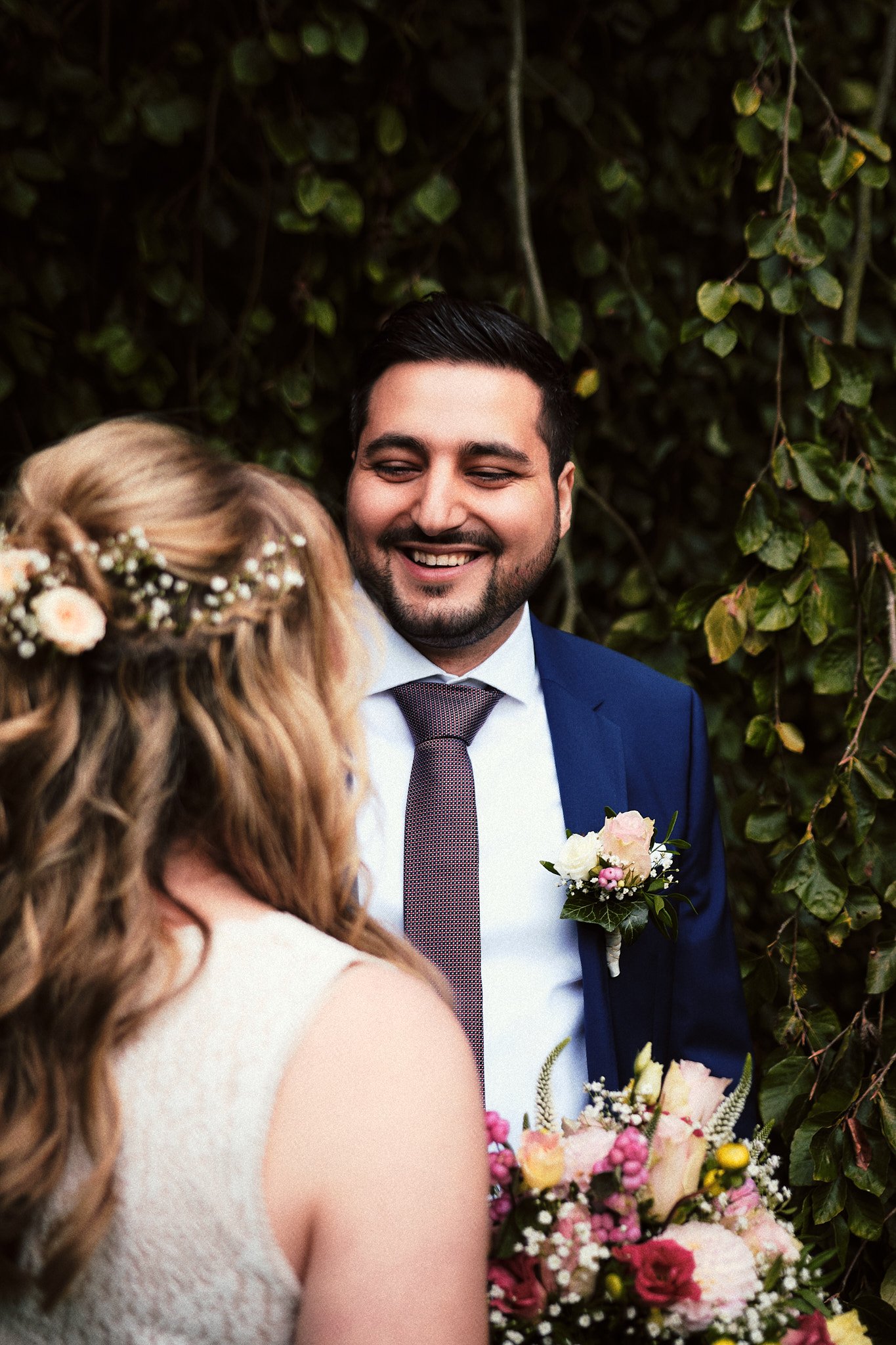 Ein lächelnder Mann in einem blauen Anzug mit Krawatte hält eine Frau mit lockigem Haar und Blumenschmuck im Haar beim Blick in ihre Richtung, umgeben von grünen Blättern und Blumen.