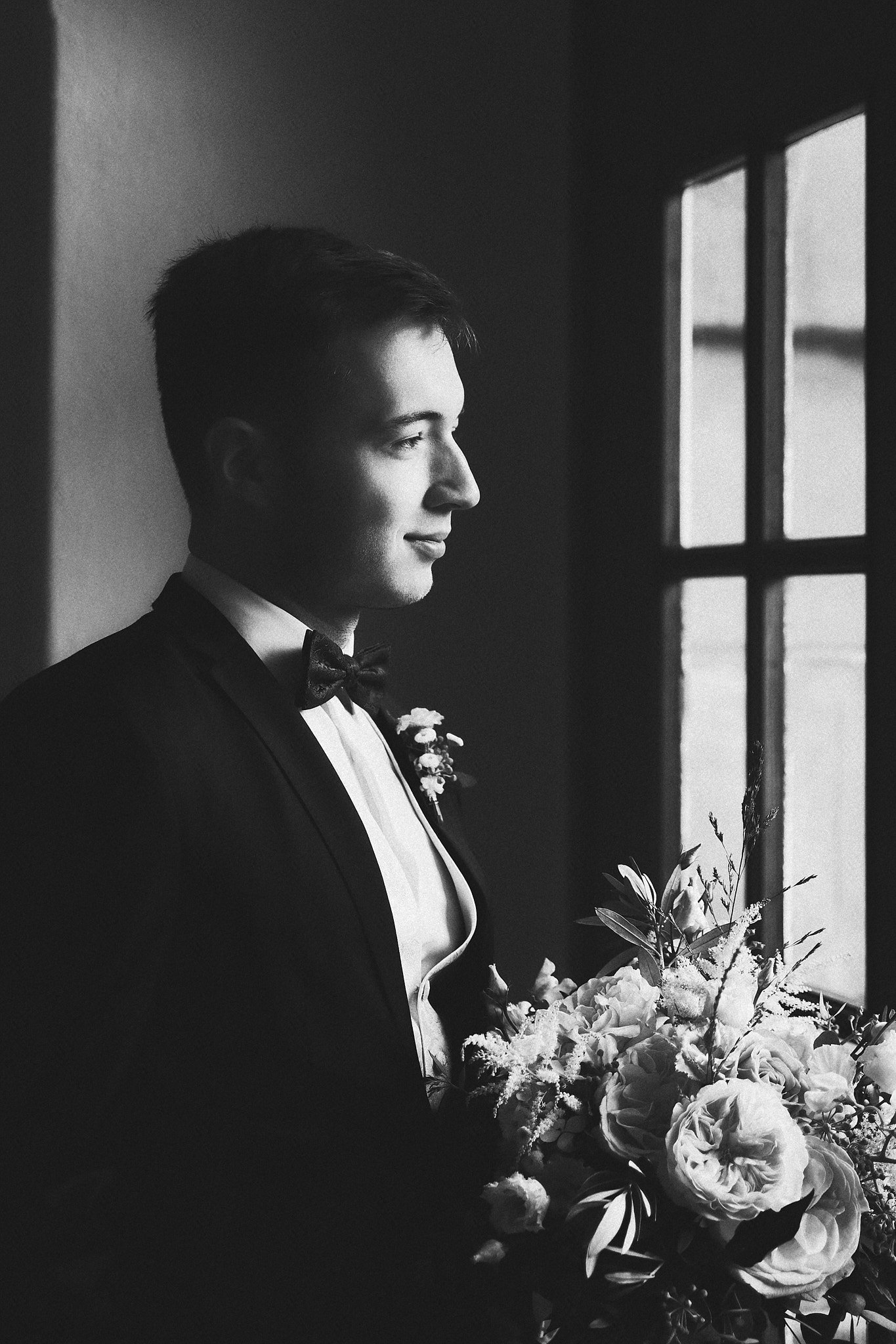 Schwarz-weiß-Foto eines Mannes in Abendkleidung mit Fliege, der ein Blumenbouquet hält, blickt zur Seite und steht neben einem Fenster.