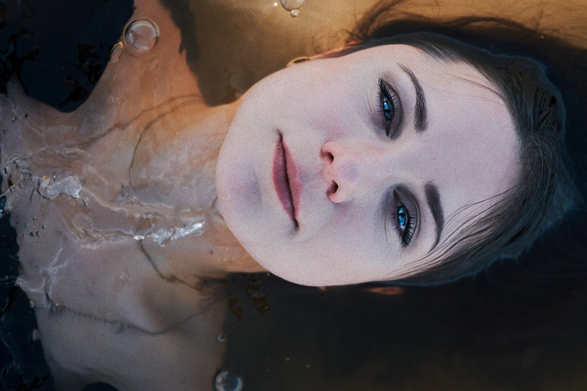 Nahaufnahme eines Schönheitsporträts einer jungen Frau mit blauen Augen, die im Wasser liegt, das ihr Gesicht umgibt.