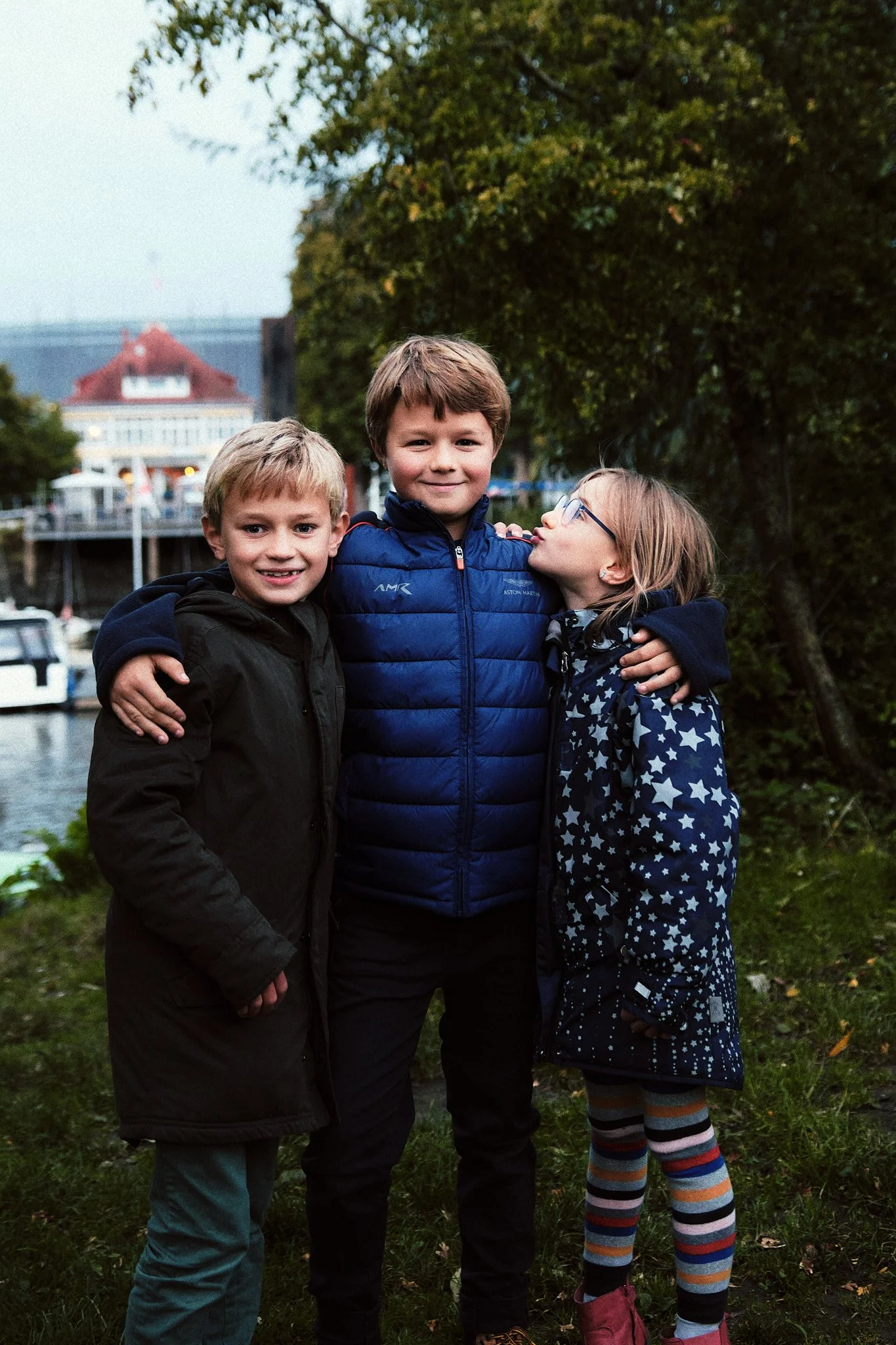 Drei Kinder umarmen sich an einem Flussufer mit Booten und Bäumen im Hintergrund.