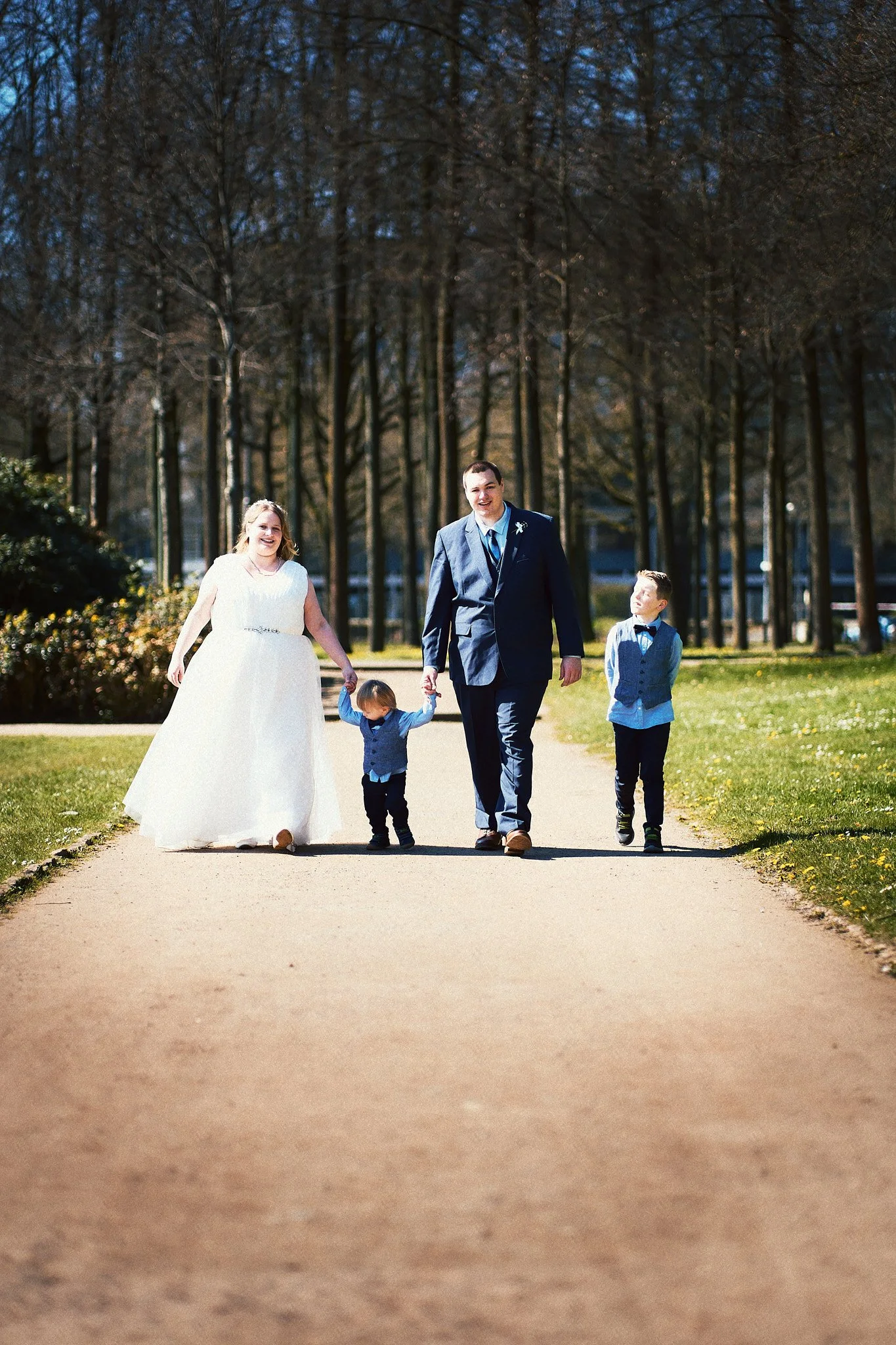 Eine Hochzeitsfamilie besteht aus einer Frau in weißem Brautkleid, einem Mann im Anzug, einem kleinen Jungen und einem älteren Jungen, die Hand in Hand auf einem Weg im Park gehen.