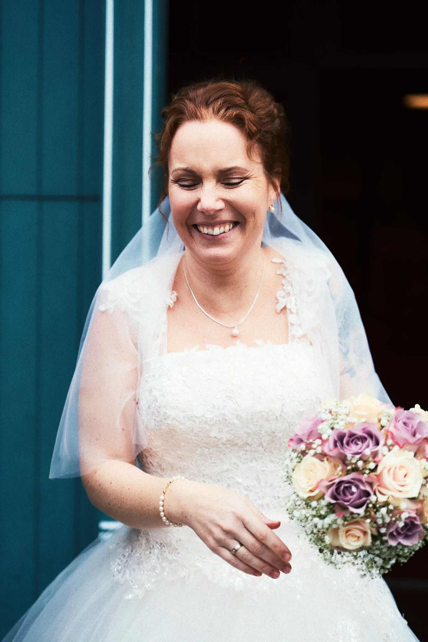 Eine lächelnde Braut in einem weißen Hochzeitskleid mit einem Brautstrauß aus rosa und lila Rosen und weißen Blumen, vor einem blauen Gebäude.