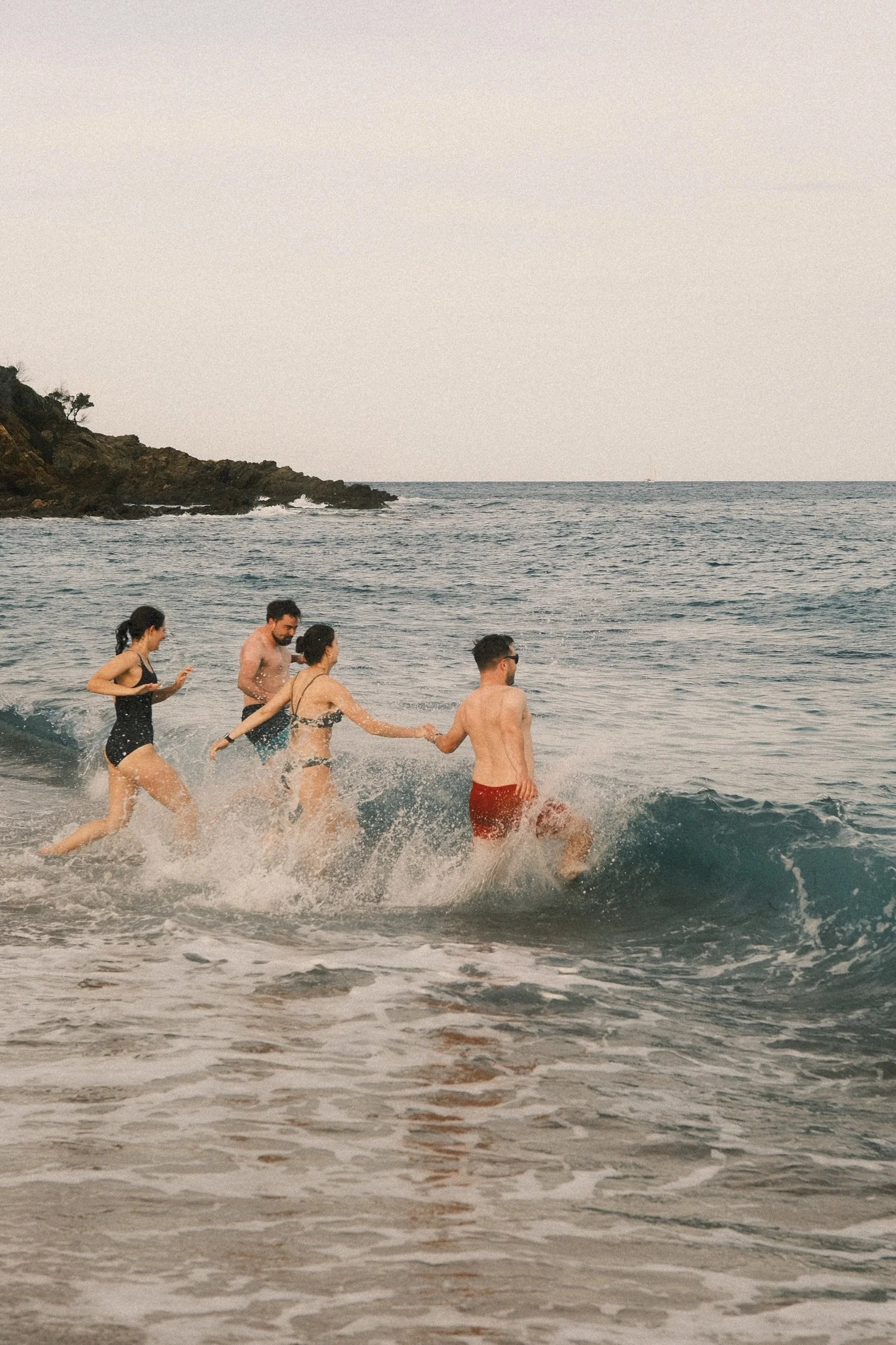 Fünf Menschen springen ins Meer, während sie am Strand laufen, mit einer Klippe im Hintergrund und einer ruhigen See.