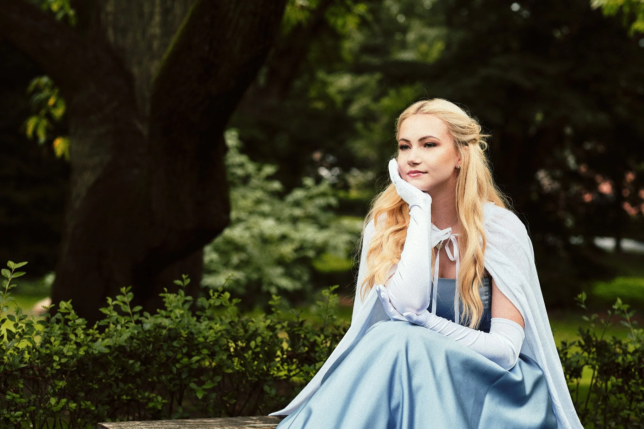 Eine Frau in einem blauen Kleid, weißen Handschuhen und einem weißen Umhang sitzt nachdenklich auf einer Bank in einem Park mit vielen Bäumen im Hintergrund.