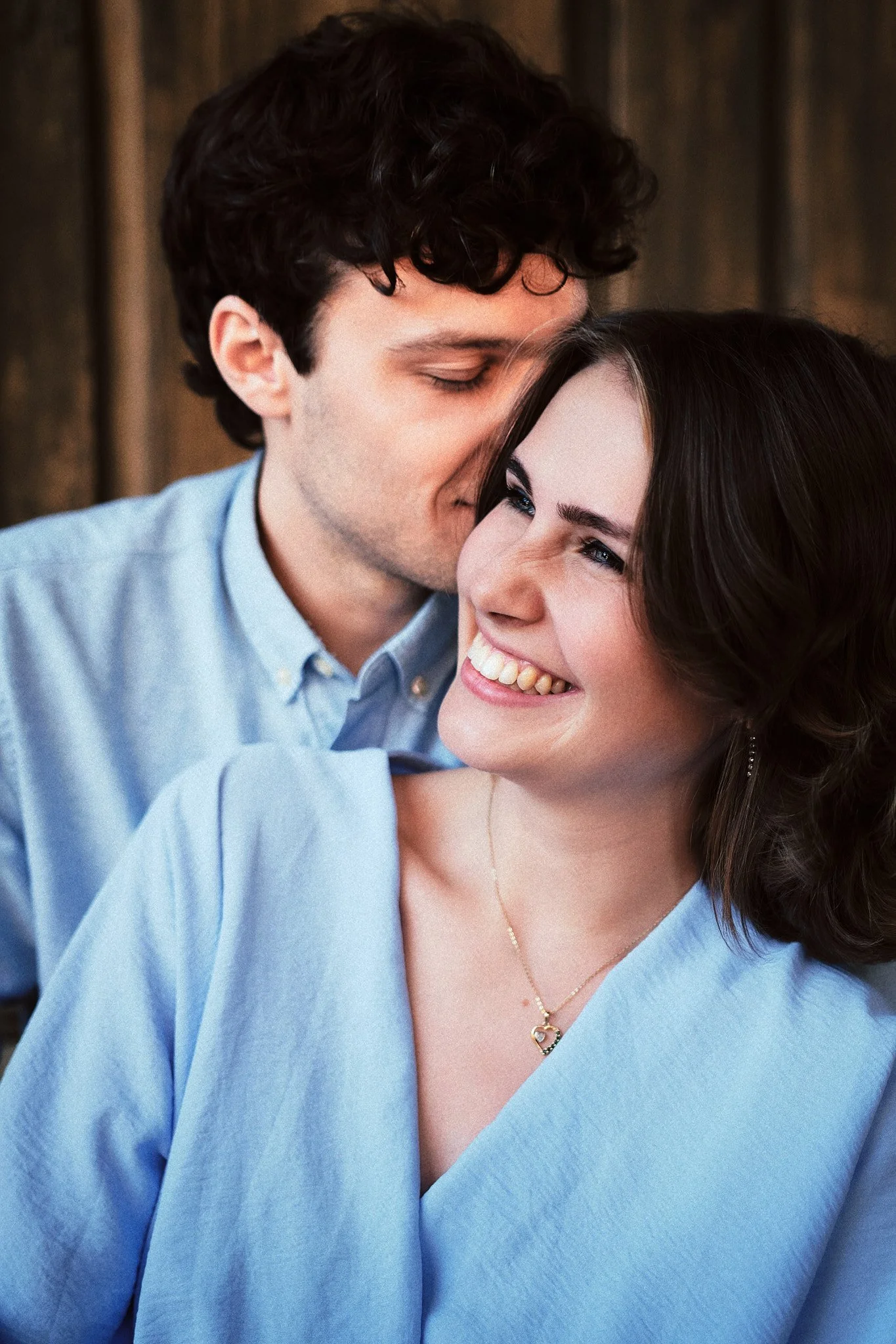 Ein junges Paar, das sich liebevoll küsst und lacht, vor einer Holzwand.