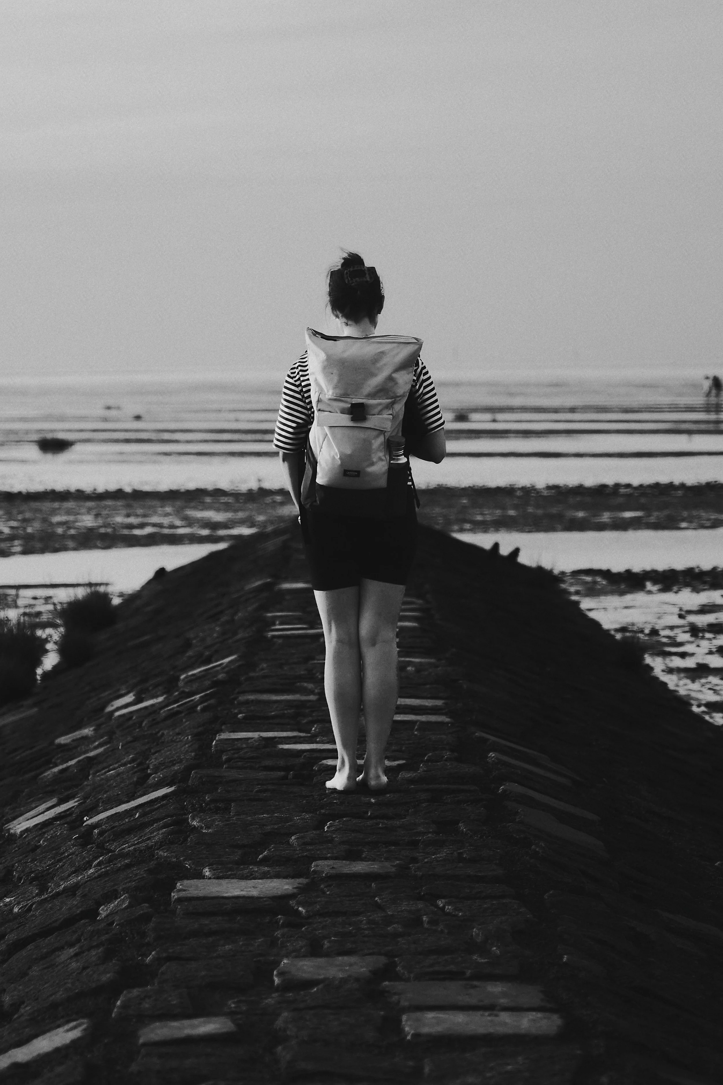 Eine Frau mit Rucksack läuft auf einem Steg an einem Meer oder einem See im Sonnenuntergang oder bei Dämmerung, im Schwarz-Weiß-Format.